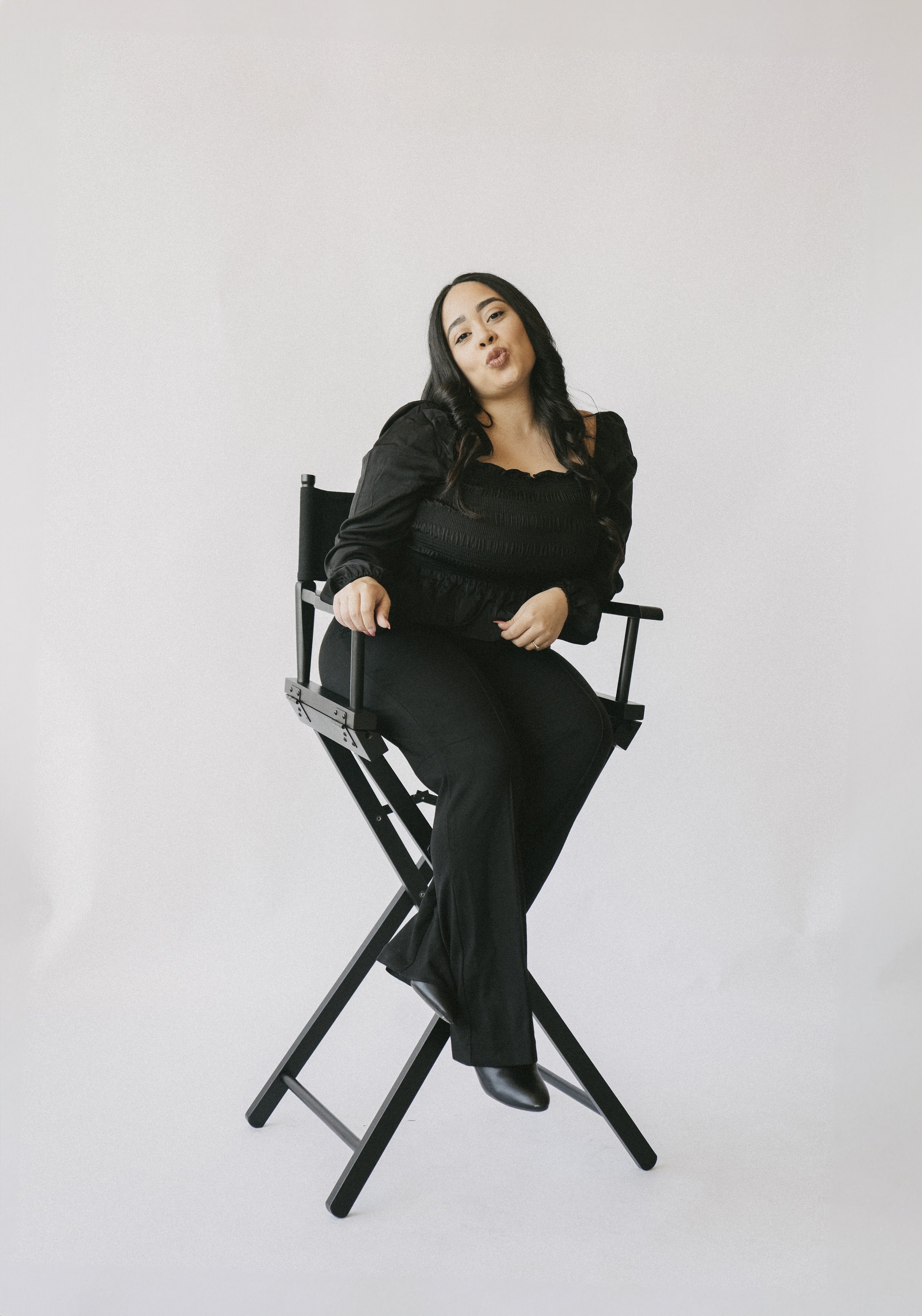 A woman with long dark hair wearing an all-black outfit posing while seated on a director's chair against a plain white background.
