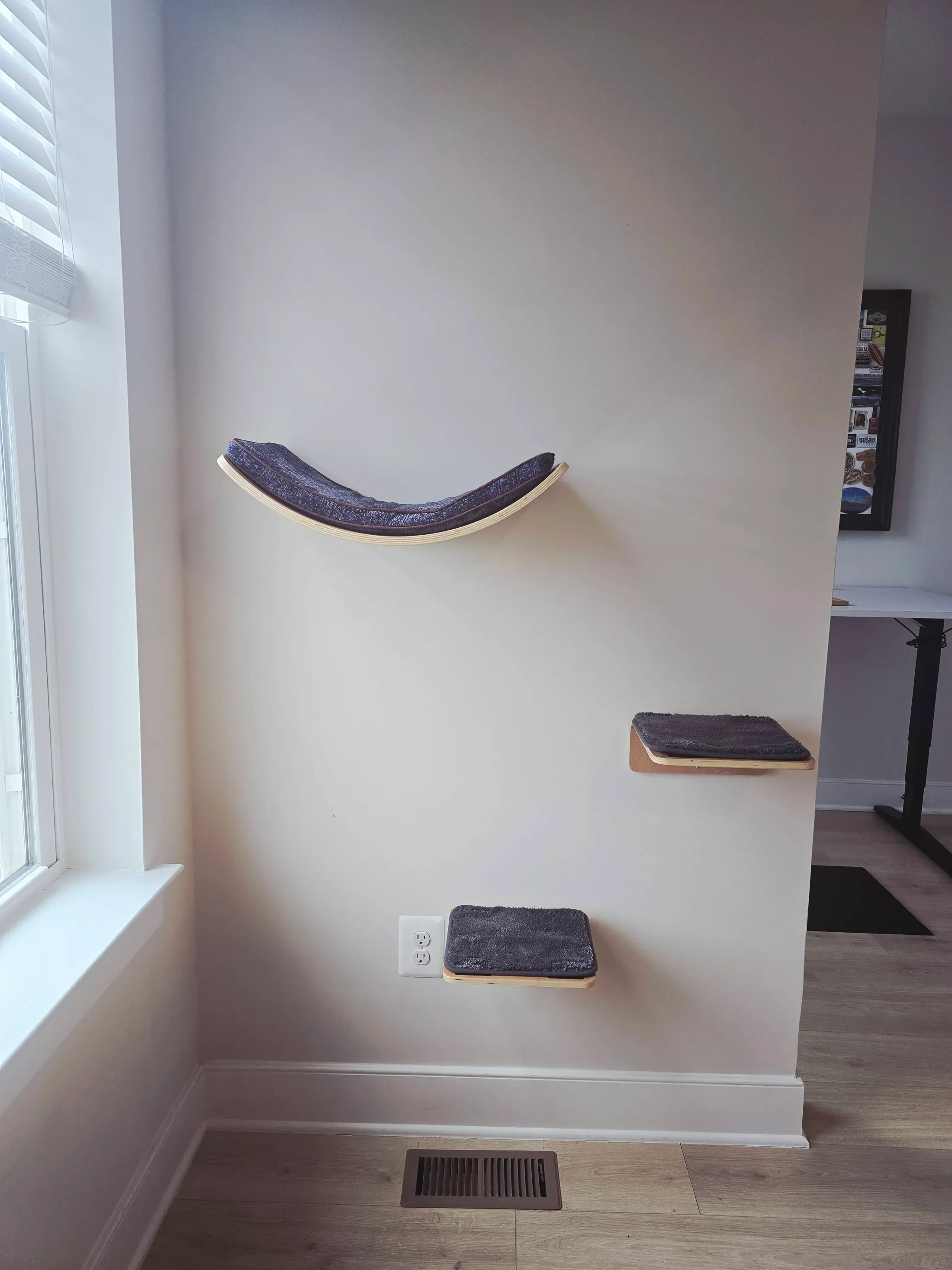 A wall with three small, black, textured cat shelves installed at different heights, a window with white blinds to the left, a white wall outlet below the middle shelf, and a floor vent at the bottom of the wall. A small black mat is placed on a wooden platform against the wall.