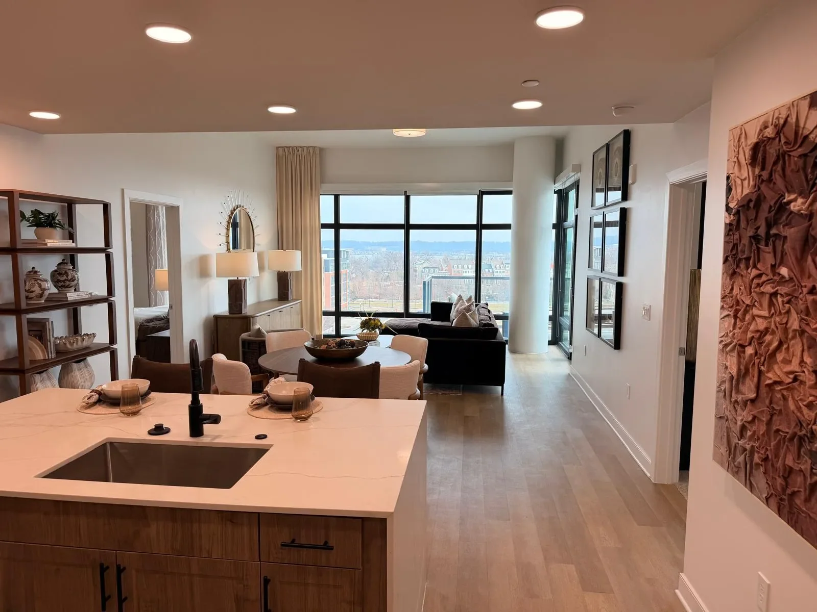 Modern open-concept living room and kitchen with a view of the city through large windows, featuring a dining table, black sofa, abstract wall art, and a storage shelf.