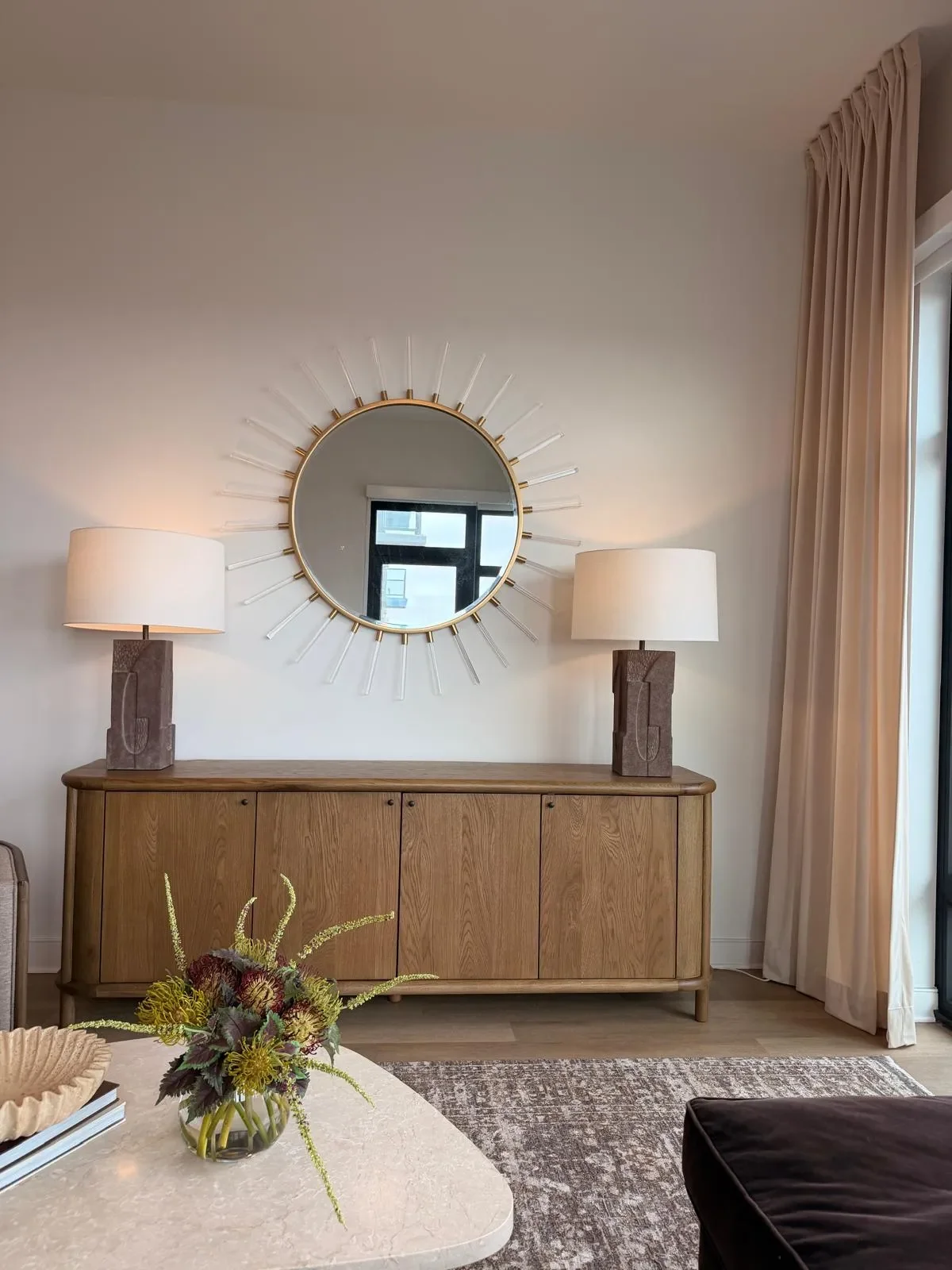 Living room with wooden sideboard, round sunburst mirror, two table lamps, beige curtains, and floral centerpiece on table.