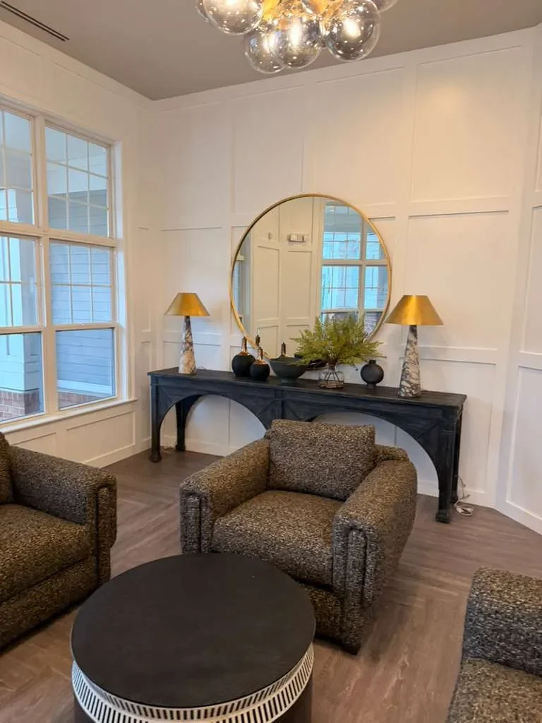Living room with two patterned armchairs, a black round coffee table, a black console table with two gold table lamps, a large round mirror, and a fireplace mantle with decorative vases and greenery, near large windows.
