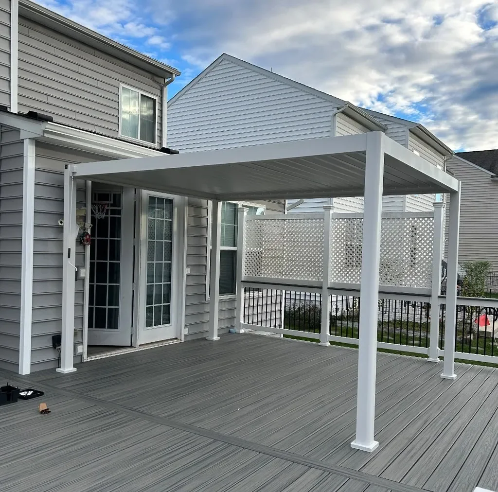 Backyard deck with a white pergola attached to the house, with siding and a sliding glass door, surrounded by other houses and a partially cloudy sky.