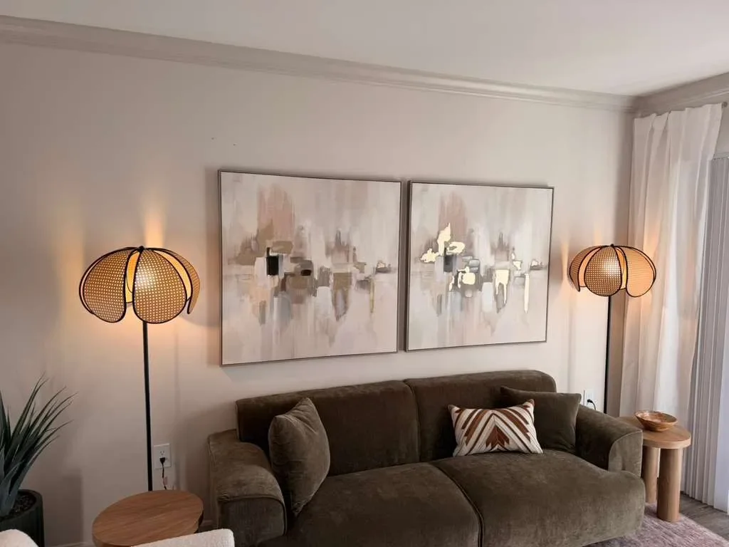Living room with a brown sofa, two abstract paintings, two floor lamps with round, Vespa-like shades, a side table with a bowl, and a curtain covering a window or sliding door.