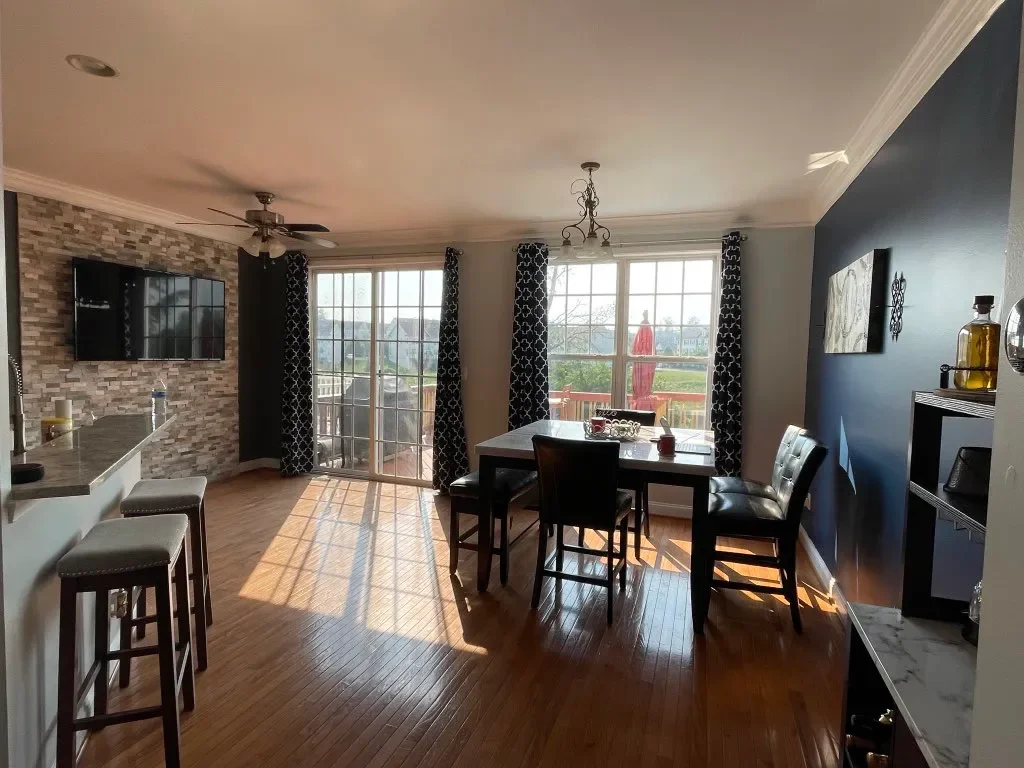 Dining area with a table and chairs by large windows, a sliding glass door leading to a deck, a brick accent wall with a mounted TV, hanging light fixtures, a ceiling fan, and a dark accent wall.