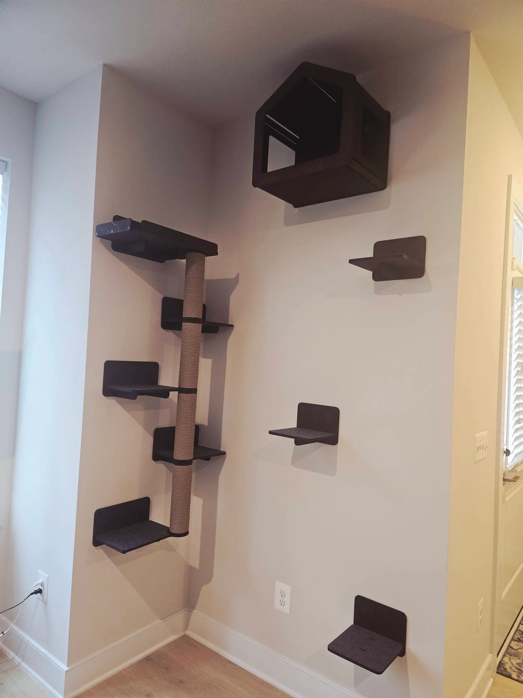 A wall with a multi-level black cat climbing structure that has platforms and a scratching post, situated in a corner near a window and electrical outlets.