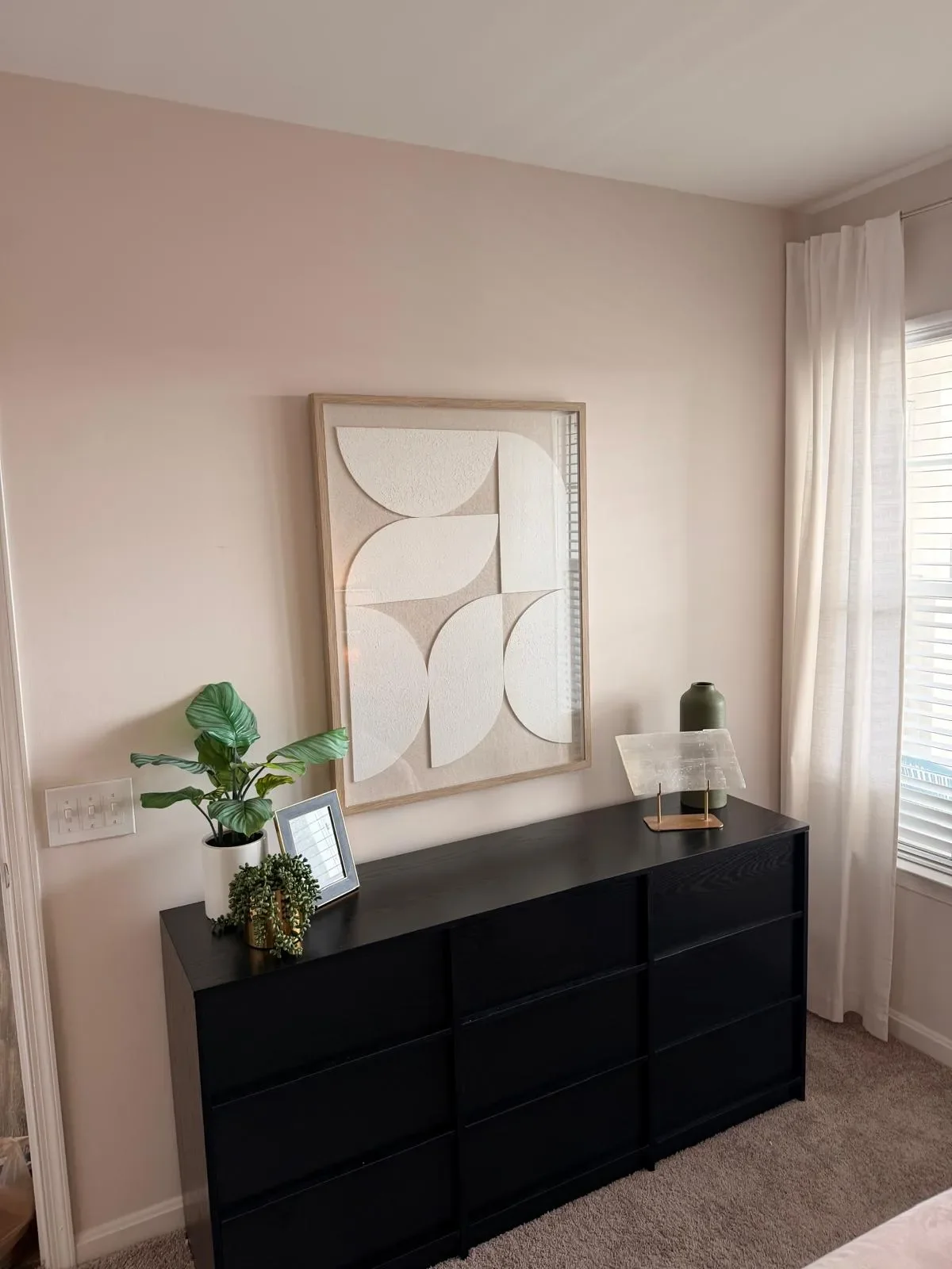 A black dresser with a potted green plant, a small mirror, a decorative, semi-transparent table lamp, and a green vase on top. A large abstract white geometric wall art hangs above the dresser. Light-colored curtains and a window are to the right.