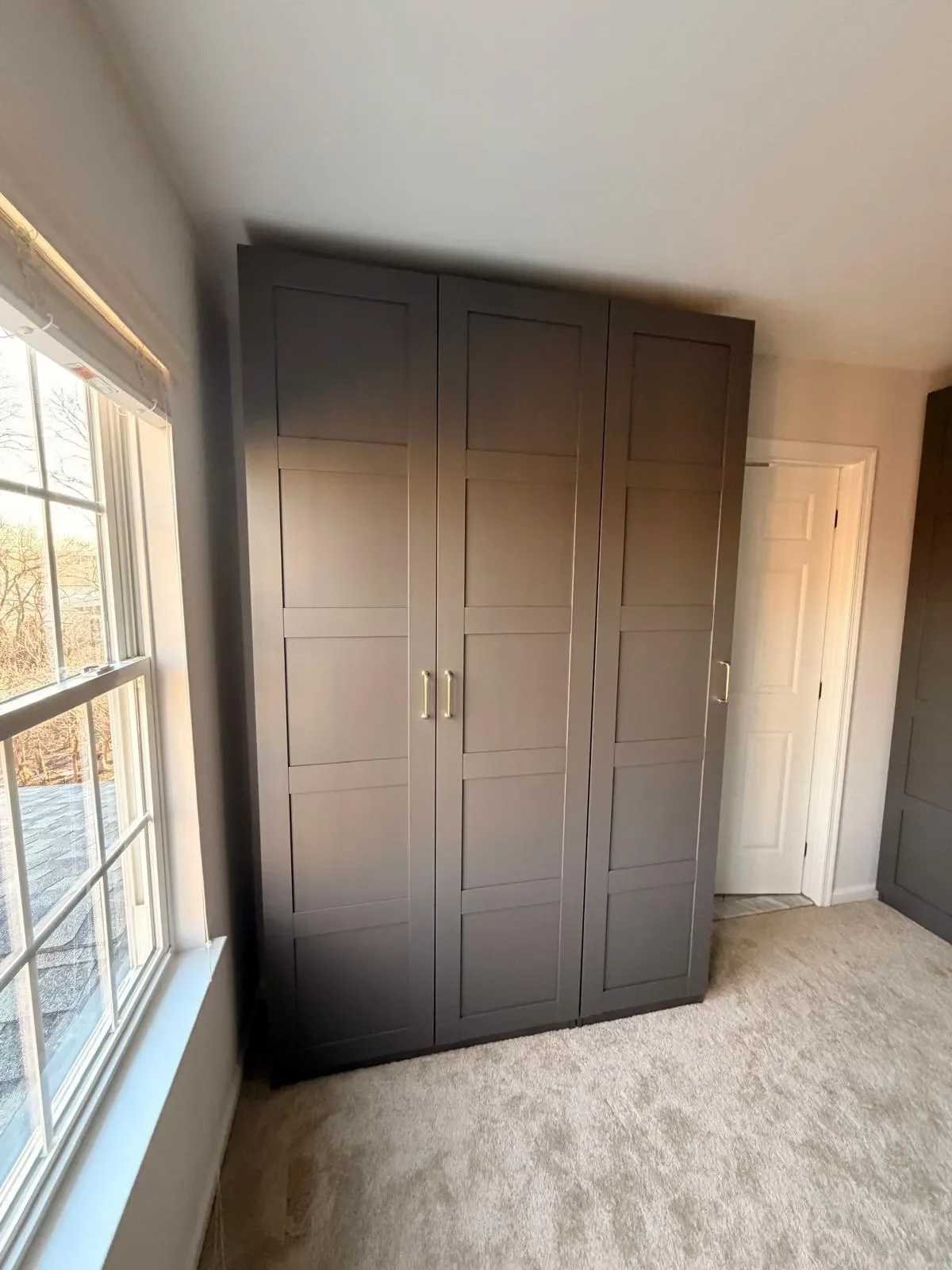 Gray built-in closet cabinet in a room with carpeted floor, positioned next to a white-framed window and a white door.
