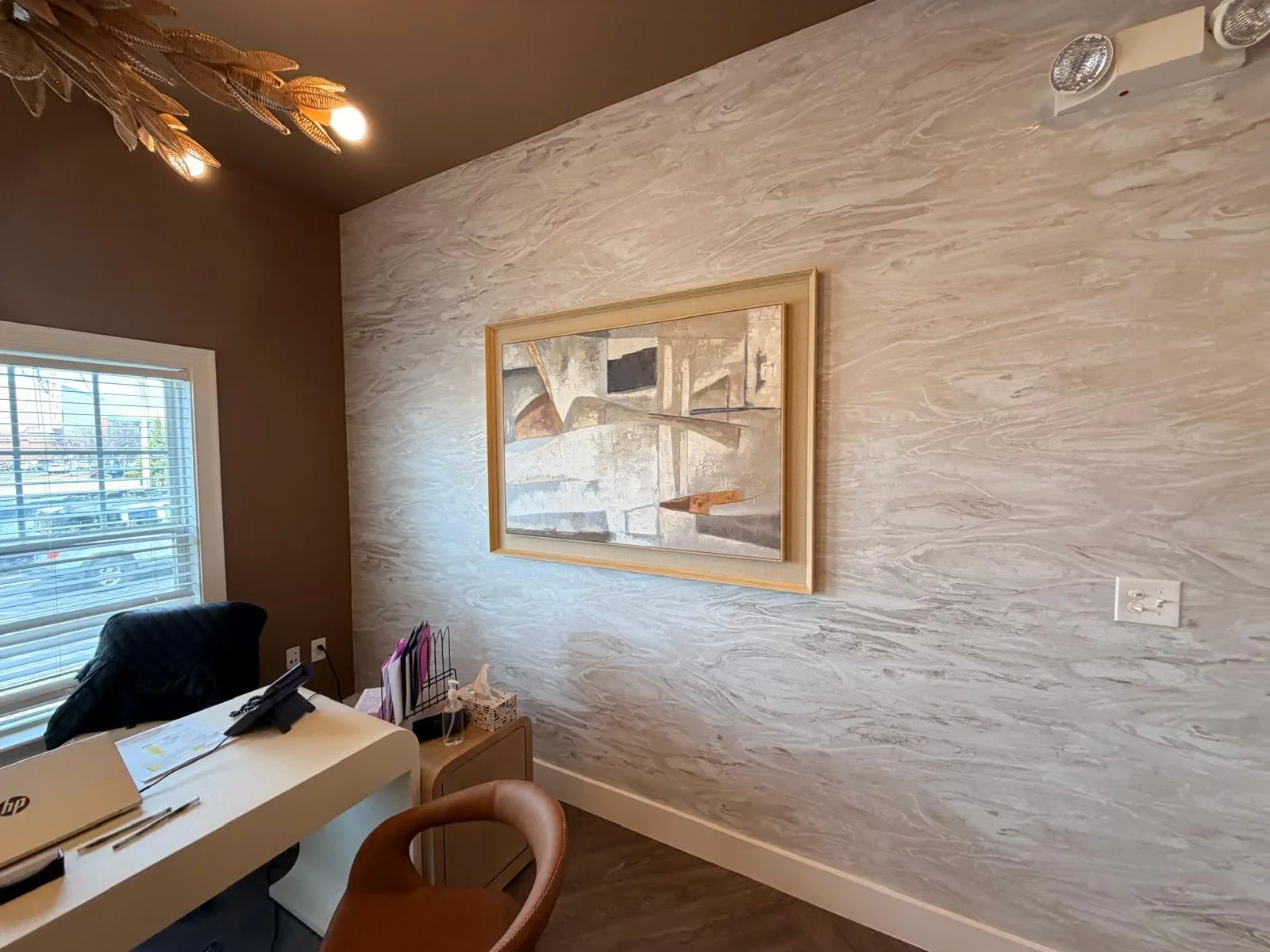 Interior of an office with a desk, black chair, window with blinds, wall art, and a textured wall.