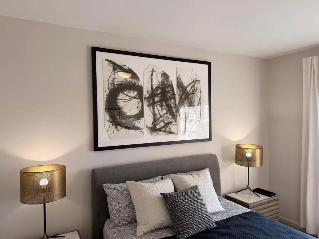A bedroom with a bed, two bedside lamps, framed abstract art above the headboard, and a window with white curtains.