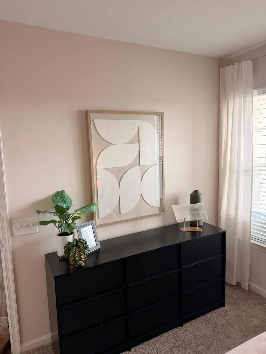 A black dresser with plants, framed artwork, a lamp, and a vase on top, positioned in front of a light-colored wall with a textured white artwork hanging above, next to a window with white curtains.