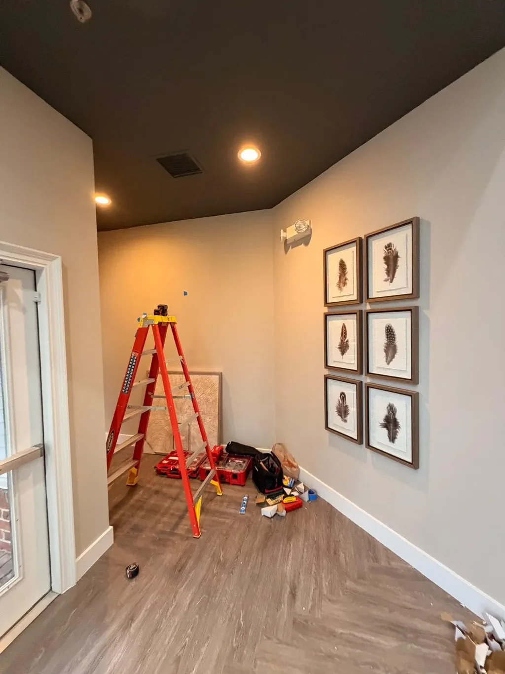 Room with a red ladder, tools, and scattered items on the floor. Framed pictures of feathers on the wall, and a partially visible door on the left.