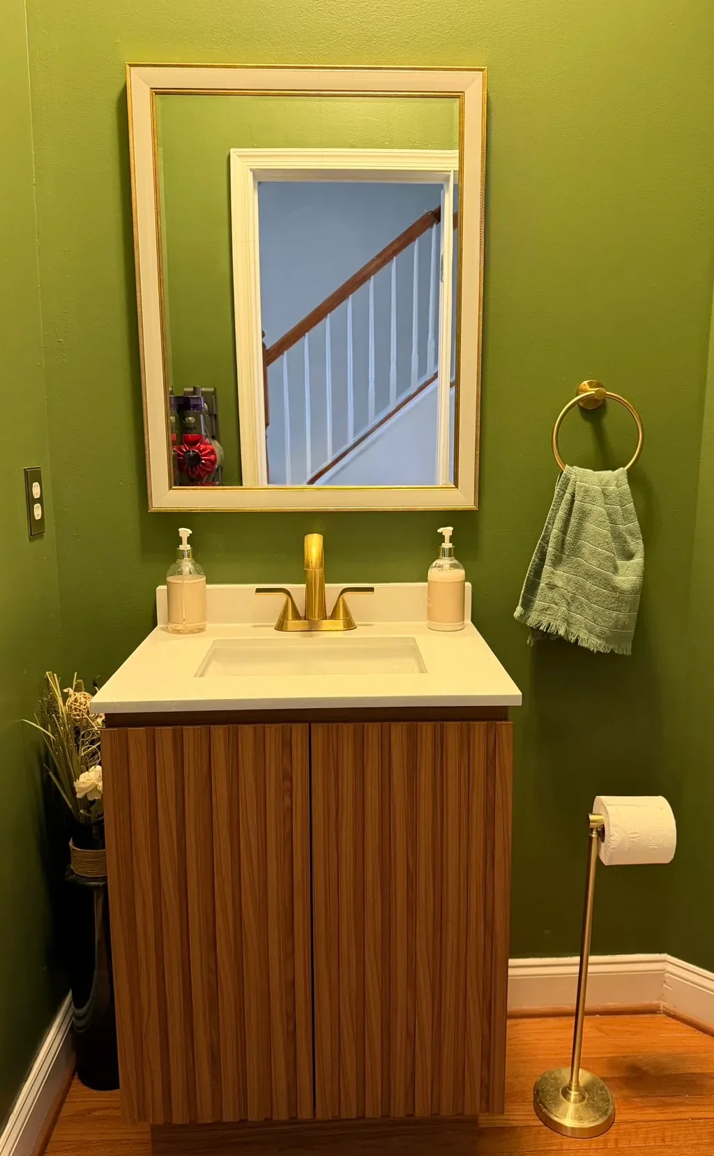 Small modern bathroom with green walls, a wooden vanity, a white countertop, and a mirror reflecting a staircase with a wooden handrail. A towel ring with a green towel and a brass toilet paper holder are on the wall. Potted plant and soap dispensers on the vanity.