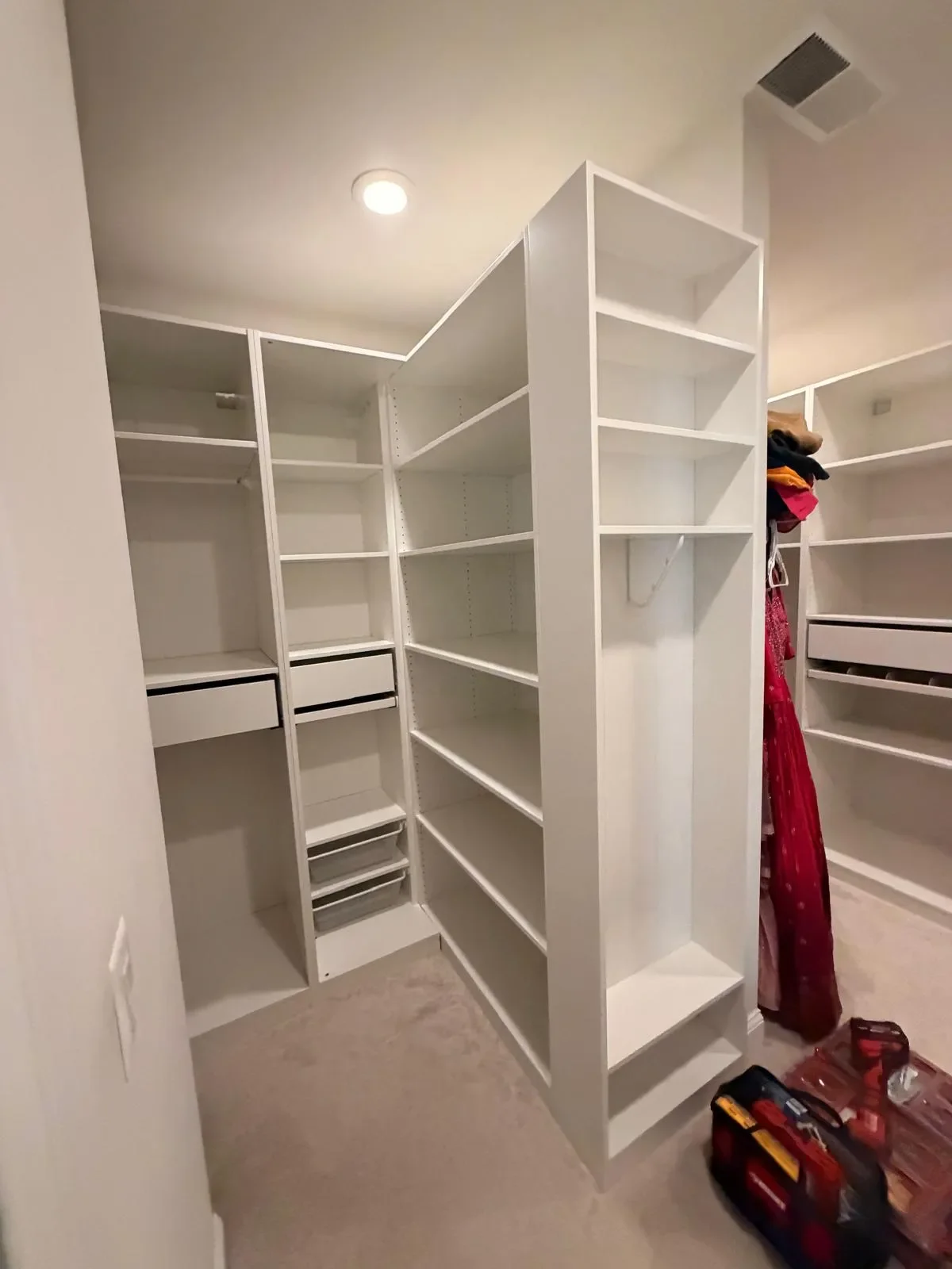 Empty white built-in closet with multiple open shelves and drawers, beige carpet, ceiling light, and scattered tools and clothing.