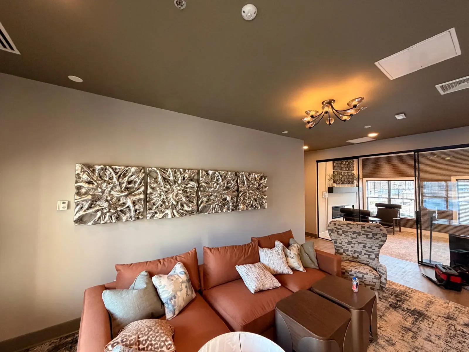 Living room with a rust-colored sectional sofa, decorative pillows, and a modern metal wall art piece. In the background, there's a glass-enclosed space with seating, a fireplace, and windows with blinds. The ceiling has a chandelier, and there's a patterned armchair near a small table.