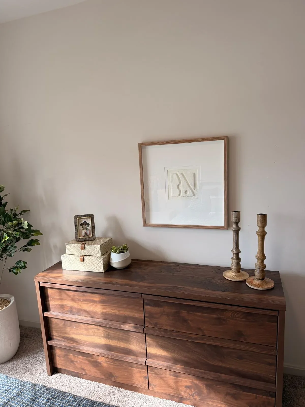 A wooden dresser with decorative items on top, including two stacked boxes, a framed picture, a white planter with succulents, and two wooden candlesticks, against a white wall with a large framed artwork.