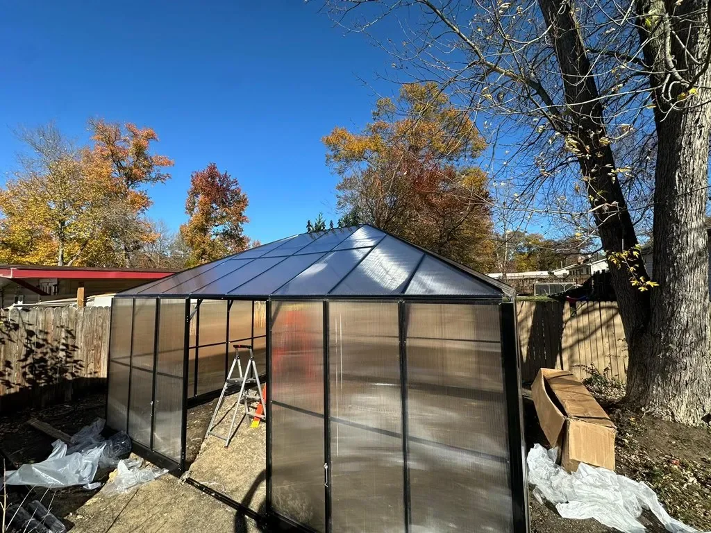 A small greenhouse with a metal frame and transparent panels in a backyard, surrounded by wooden fencing and colorful autumn trees.