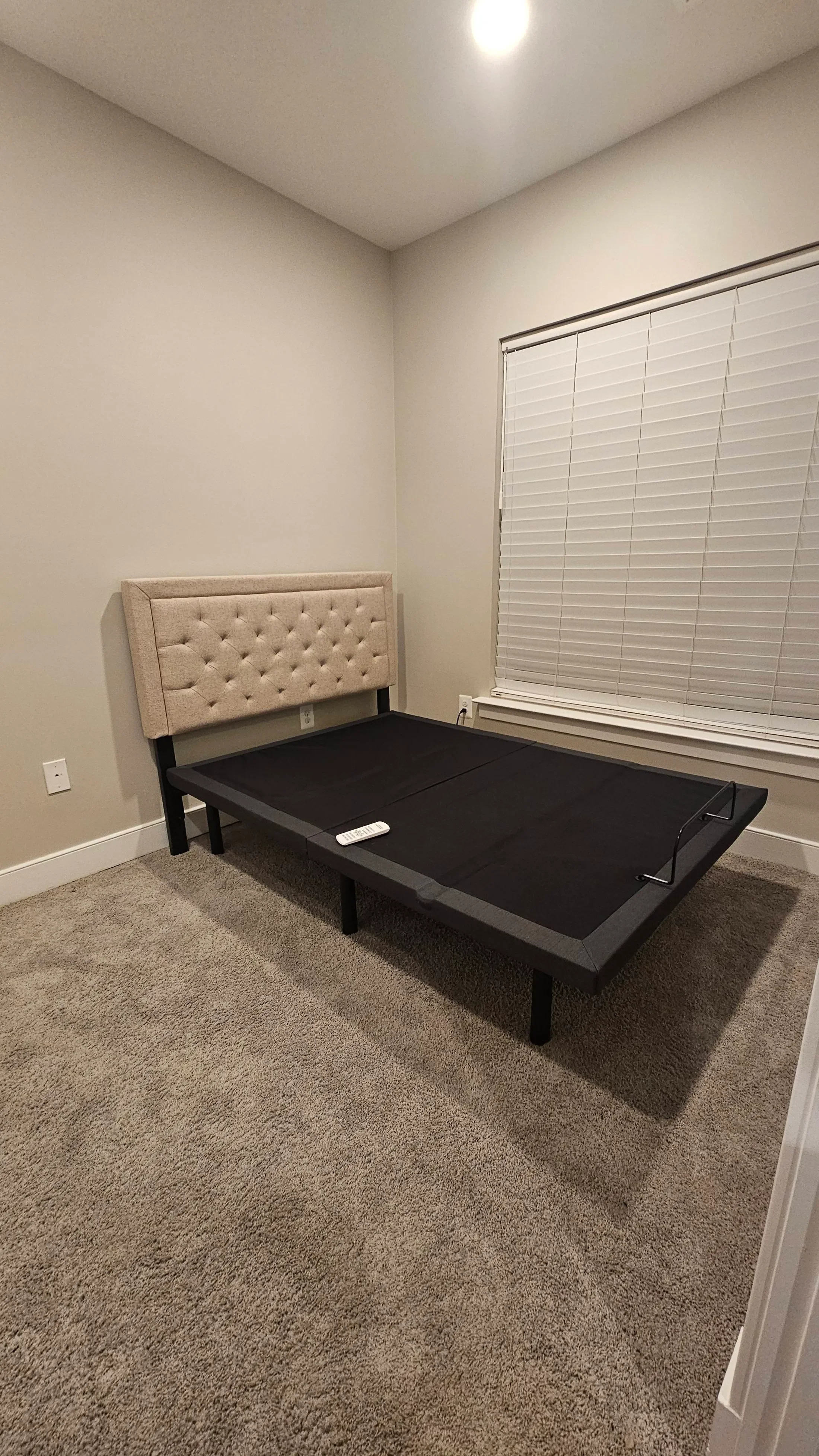 Empty bedroom with a beige upholstered headboard, a black bed frame with a mattress, a window with closed blinds, beige carpet, and a ceiling light.