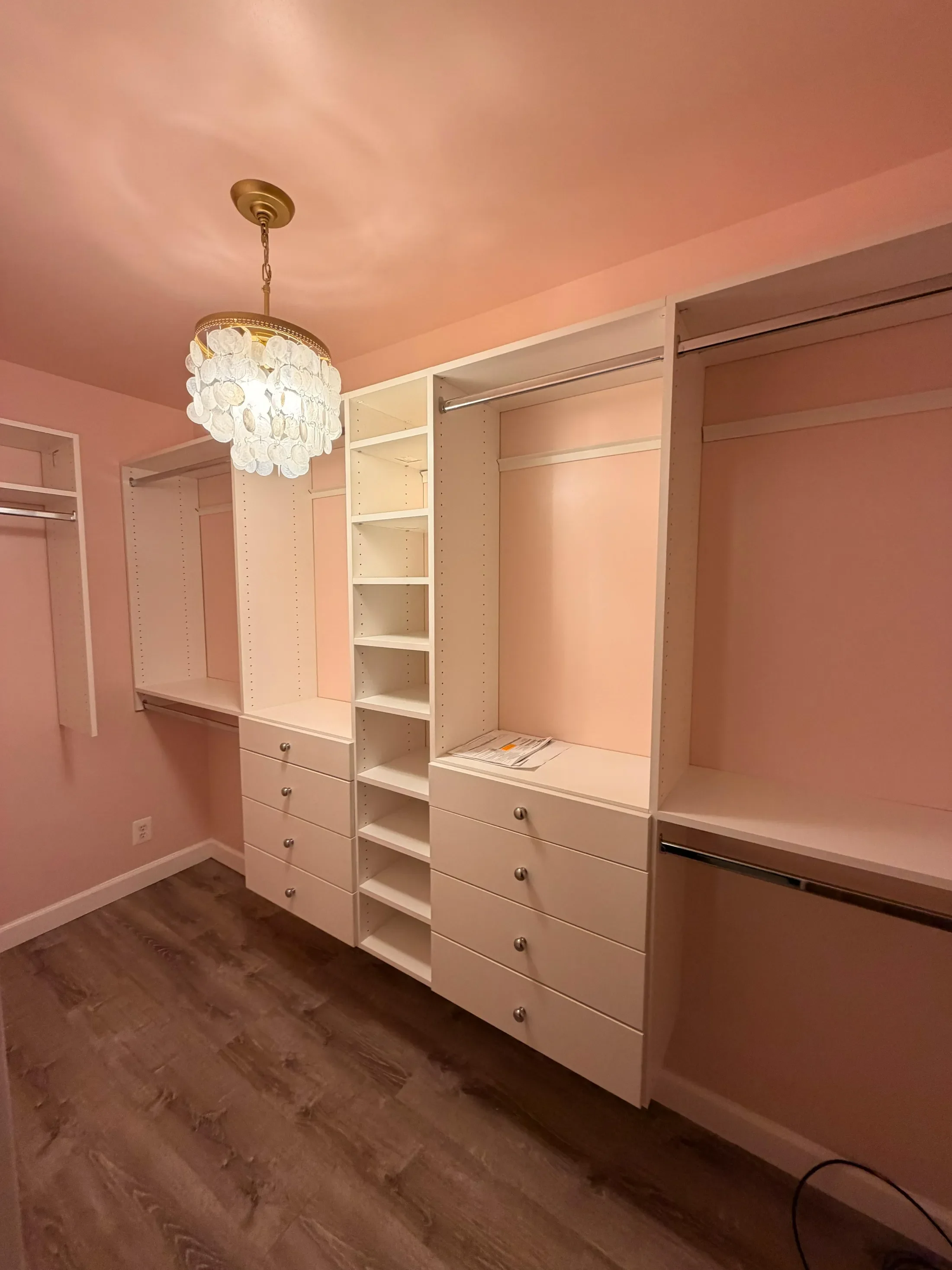 Empty white closet with open shelves and drawers, pink walls, chandelier overhead, and dark brown hardwood floors.