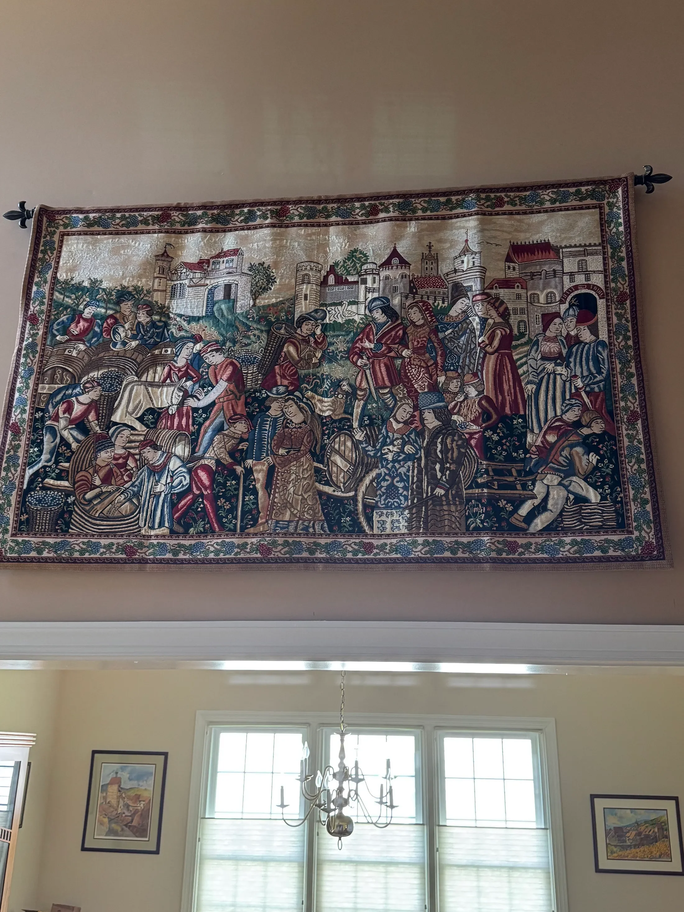 A tapestry depicting a medieval scene hangs above a large window in a room with framed artwork on the walls and a chandelier hanging from the ceiling.