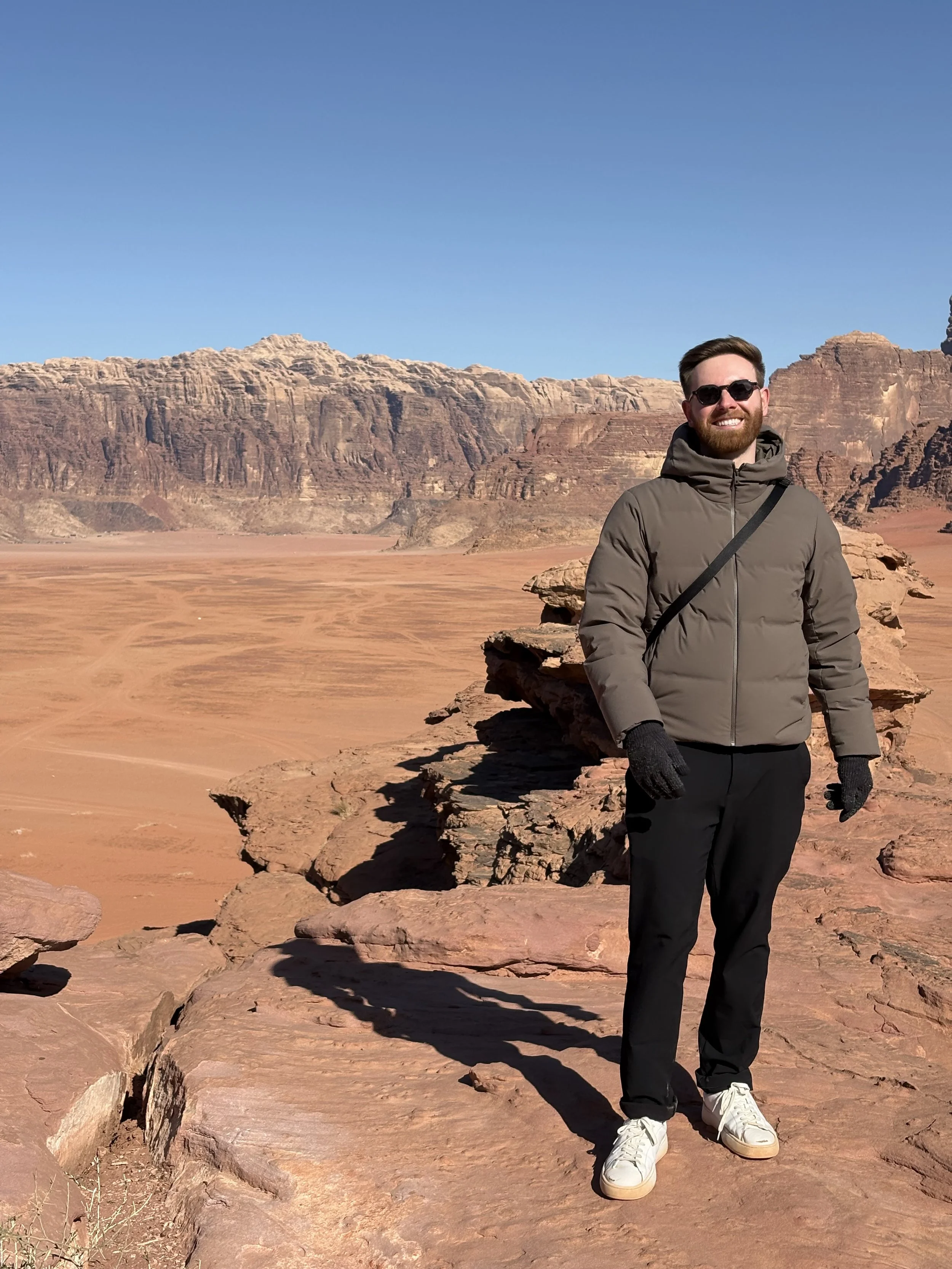 A man in a gray jacket, black pants, and white sneakers standing on red rock in a desert landscape with canyon walls and a clear blue sky