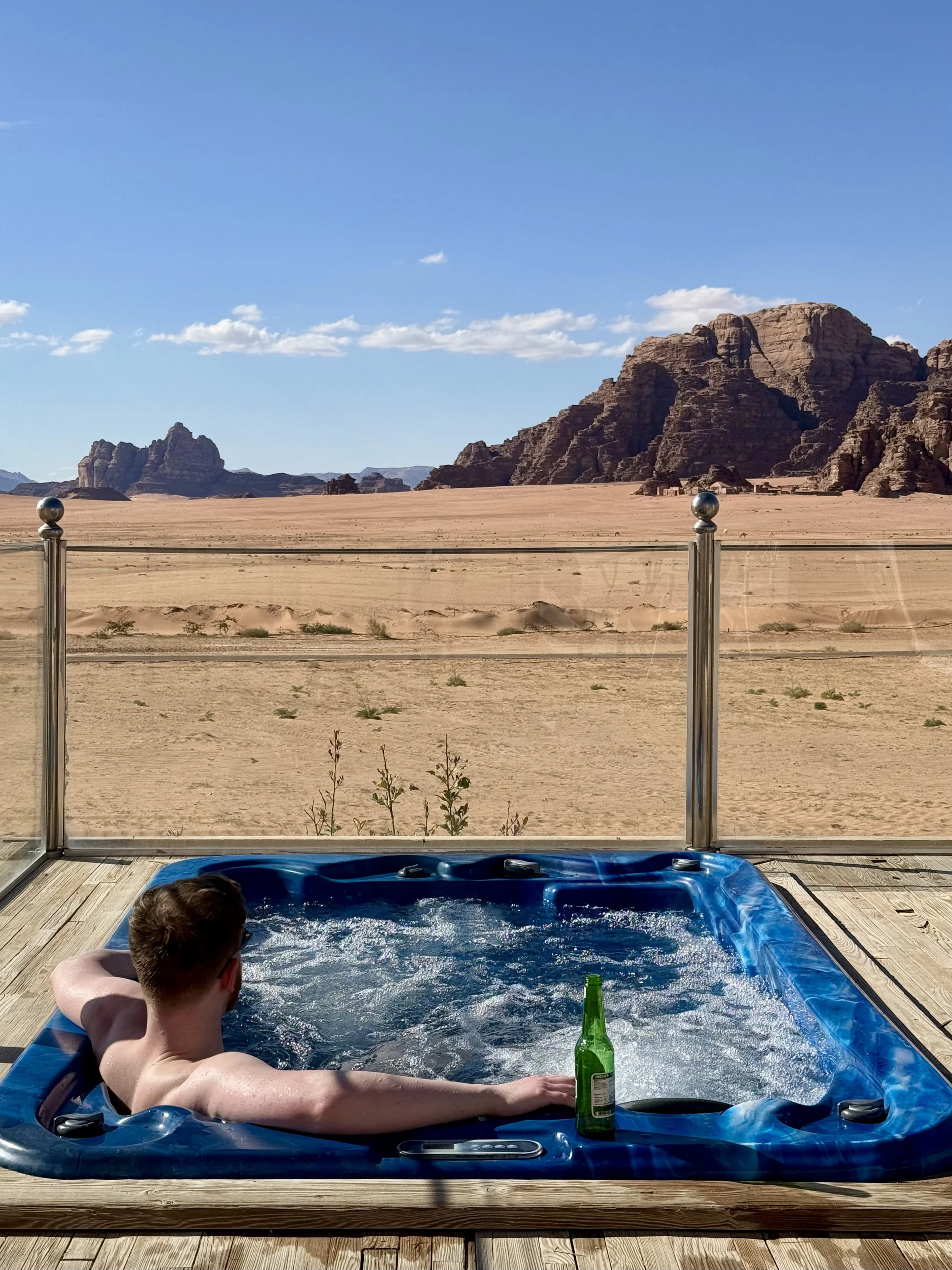 A man relaxing in a hot tub on a wooden deck with a desert landscape and rock formations in the background, holding a green bottle, under a clear blue sky.