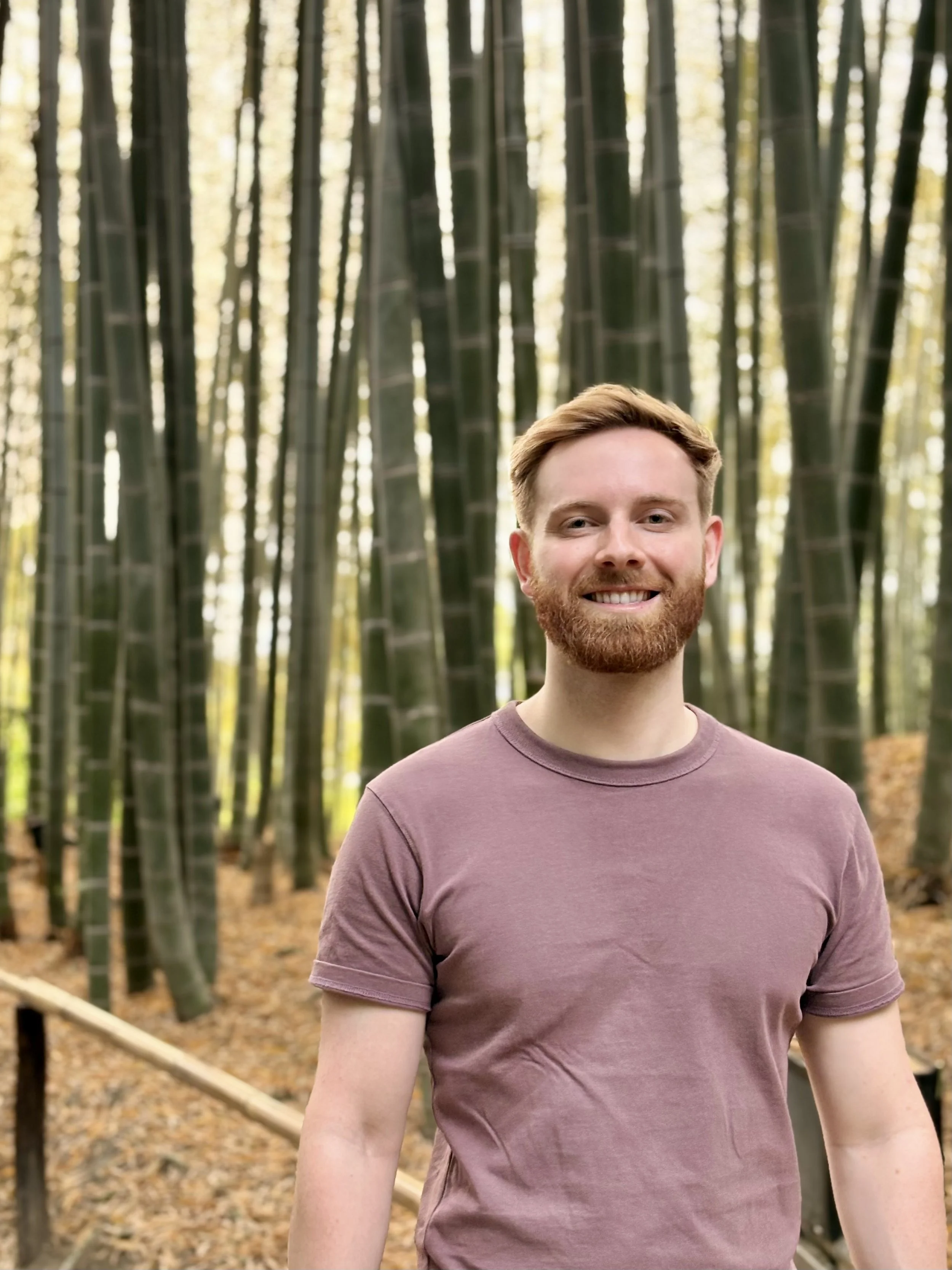 A young man with a beard and light brown hair smiling at the camera in a bamboo forest during daytime.