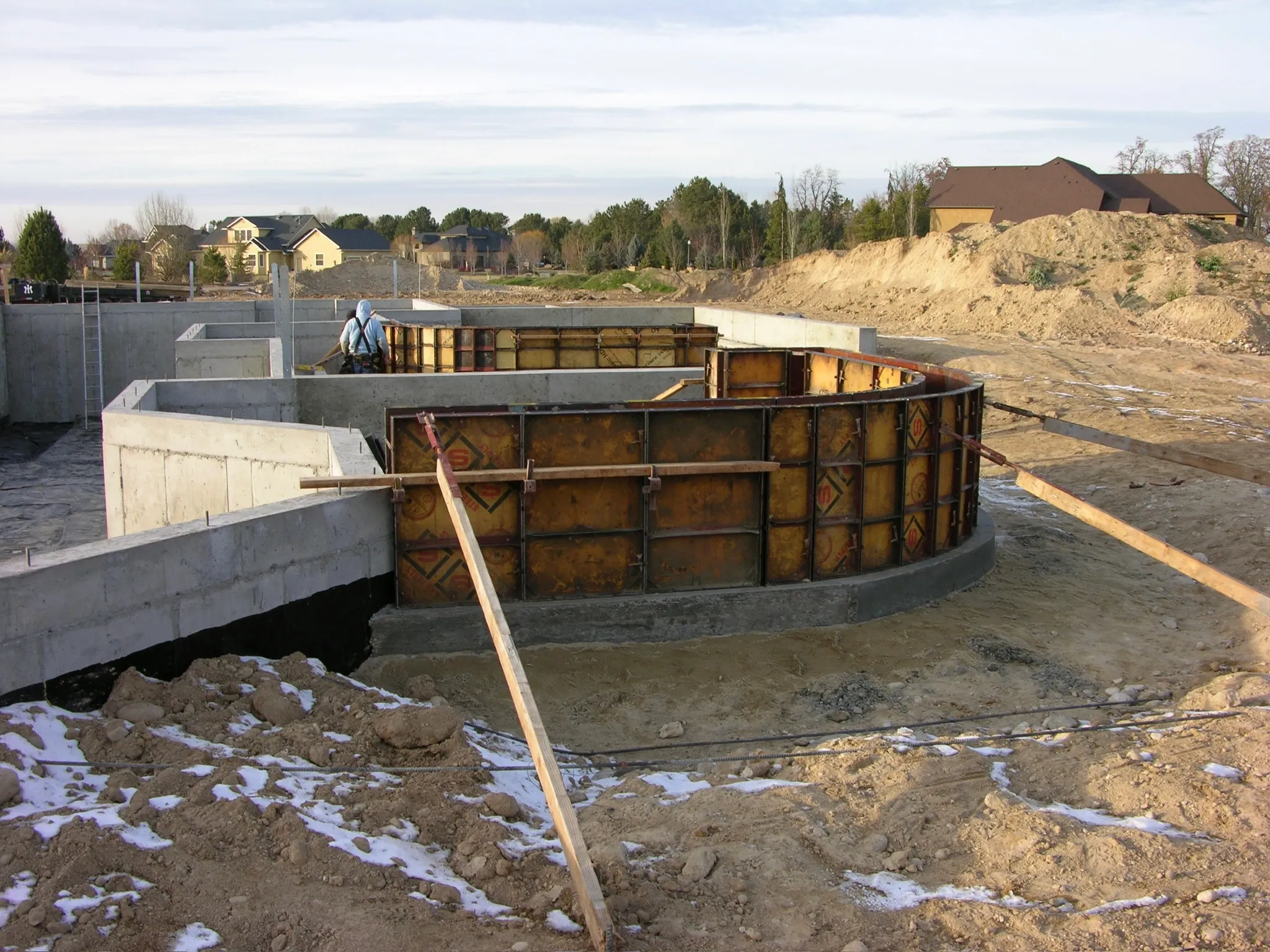 Construction site with concrete foundation walls and curved formwork for a building, workers, and construction materials in a suburban area.