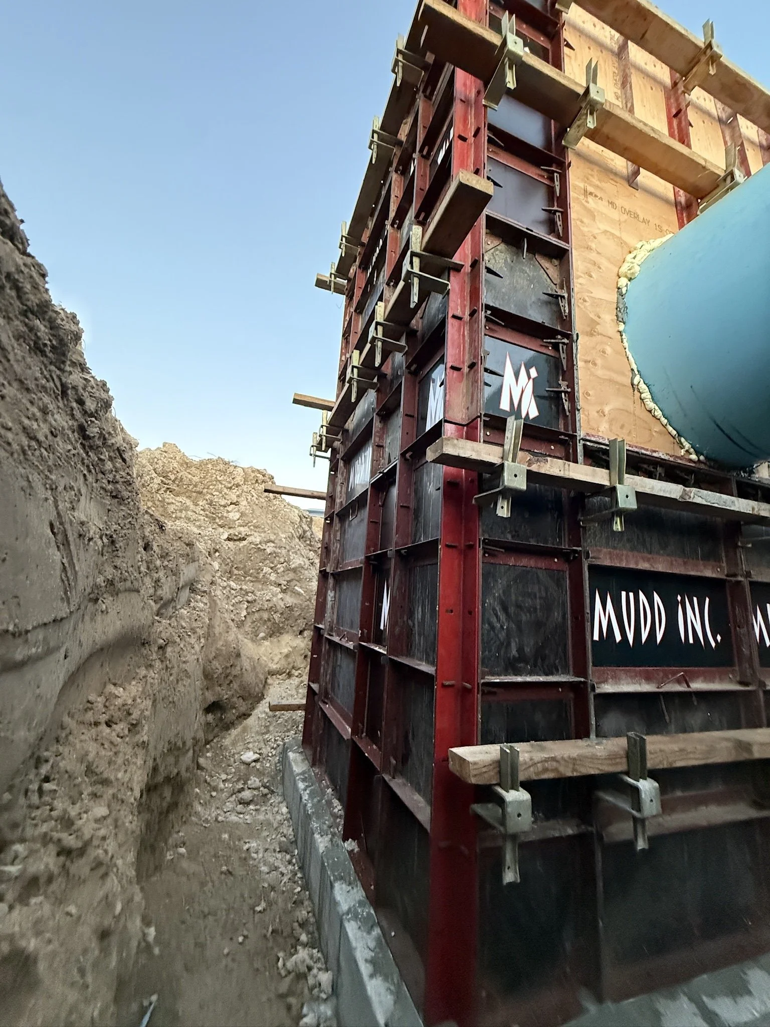 Construction site with a large partially built structure, showing red and black framework, wooden and metal supports, and a blue pipe or chute on the side, next to dug earth.