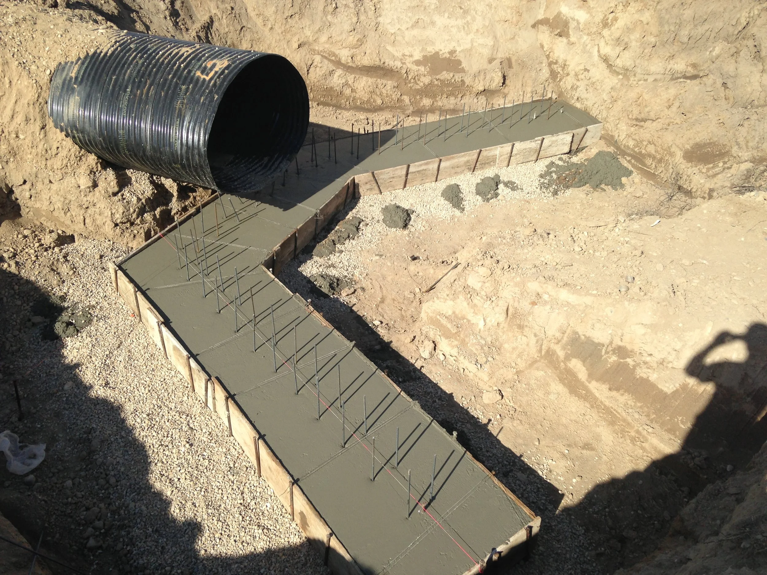Concrete foundation being poured into a wooden form with exposed rebar, with a large black corrugated pipe nearby at a construction site.
