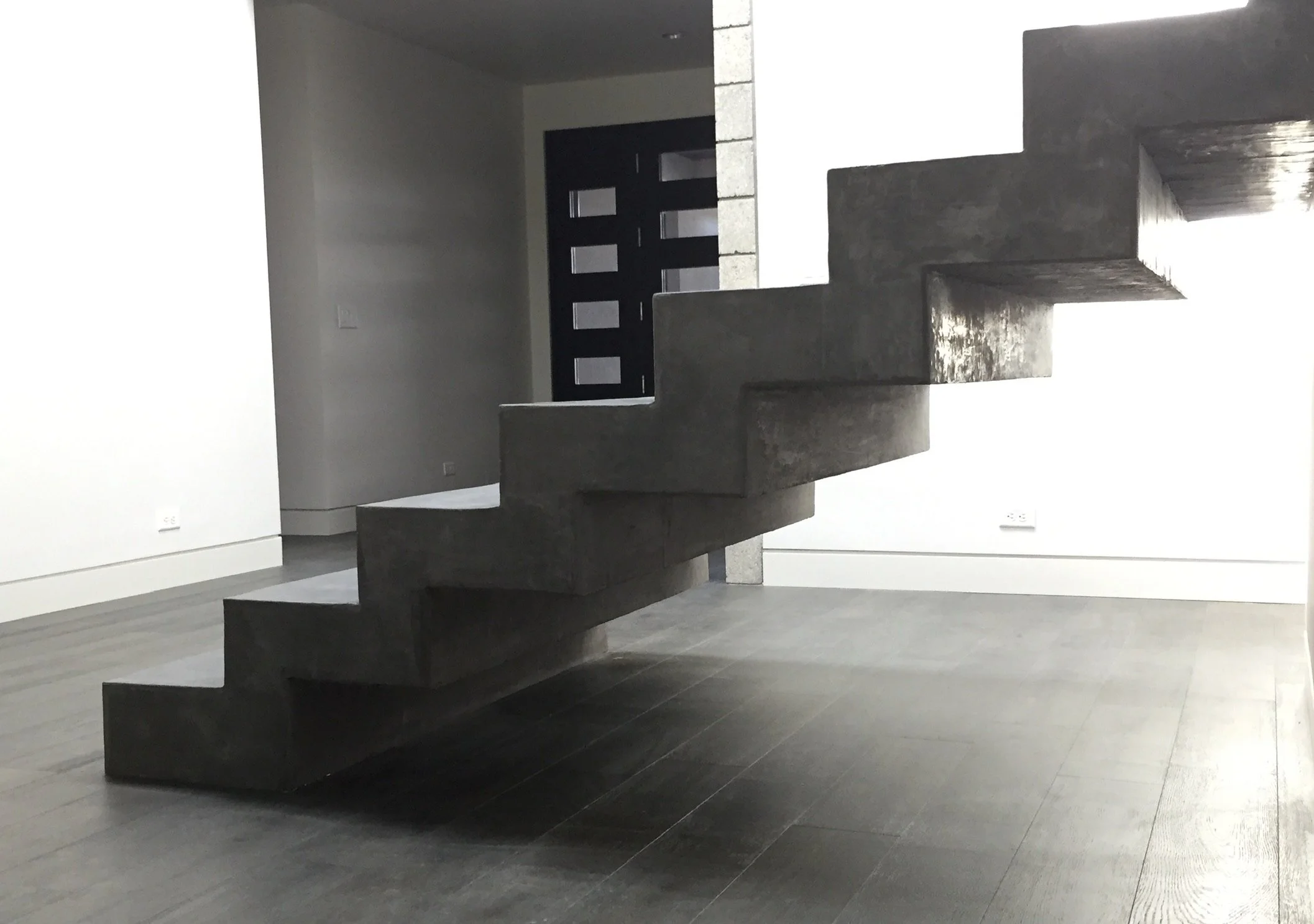 A modern, minimalist interior with a floating concrete staircase against a white wall leading to an upper level, with a black front door featuring rectangular windows in the background.