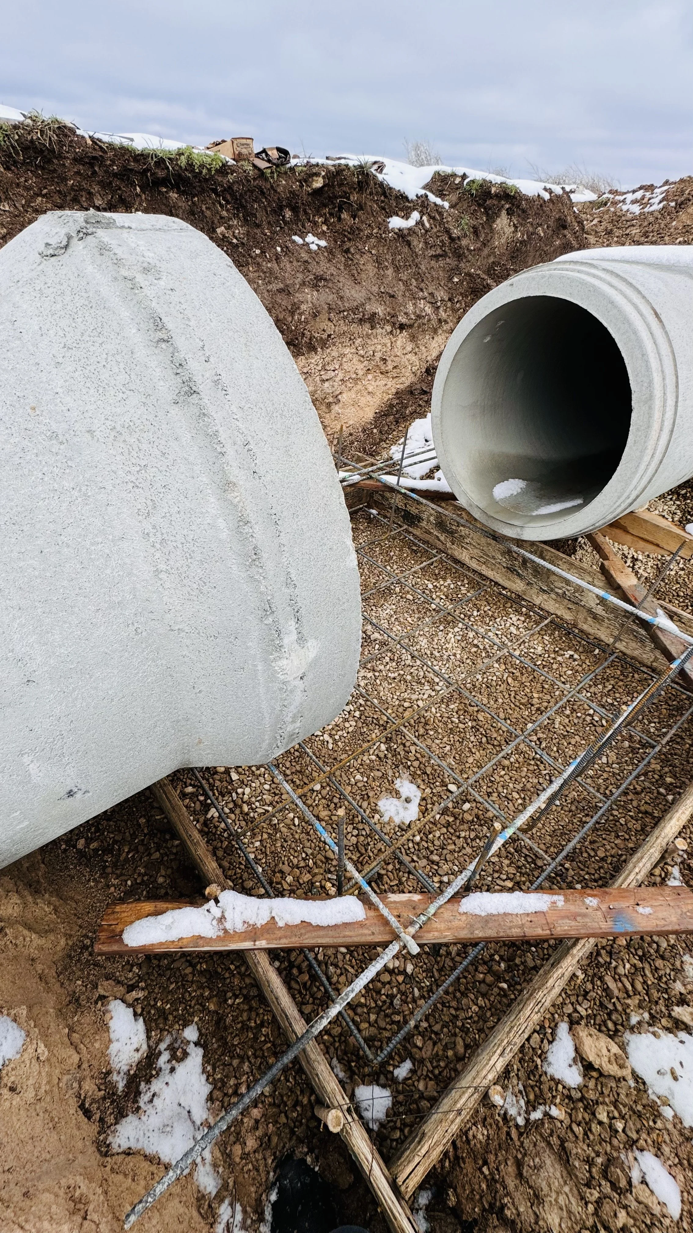 Large concrete pipes and a manhole are being installed outdoors on a construction site, with rebar and wooden supports in place, and snow on the ground.