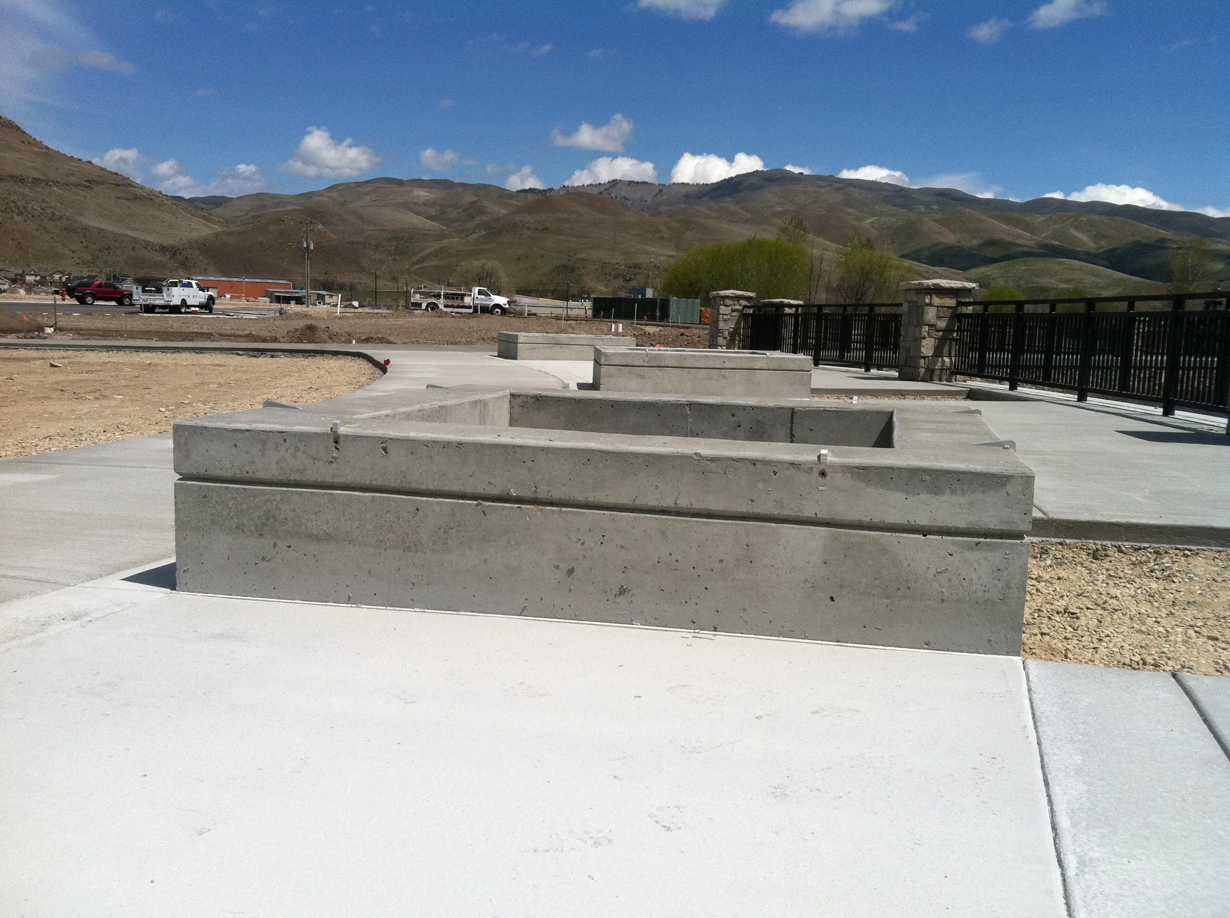Newly constructed concrete sidewalk with benches in a park, mountains in the background, and a parking lot with trucks visible in the distance.