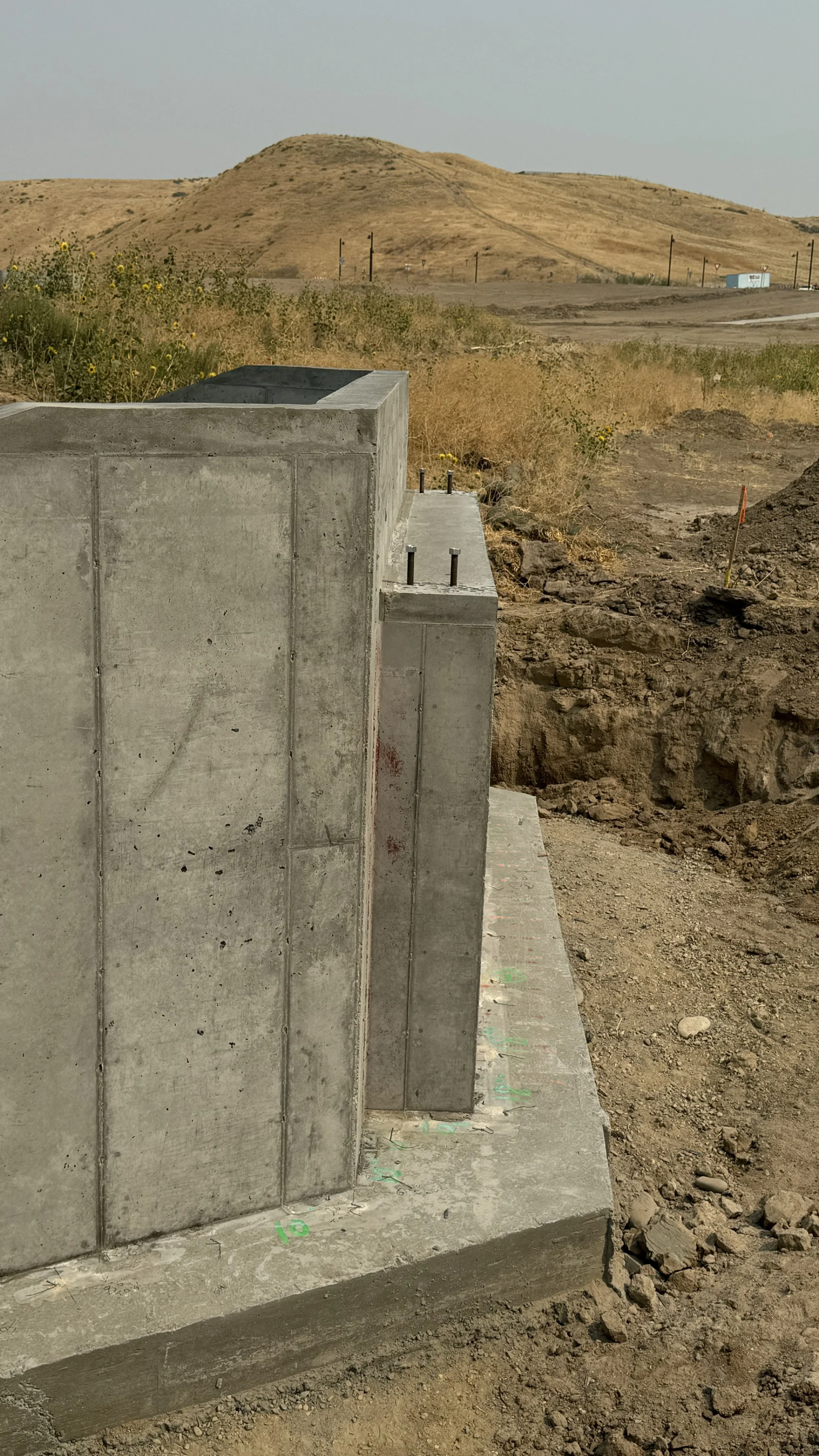 Close-up view of a concrete foundation or wall under construction at a construction site. A landscape with dry, rolling hills and sparse vegetation is visible in the background.