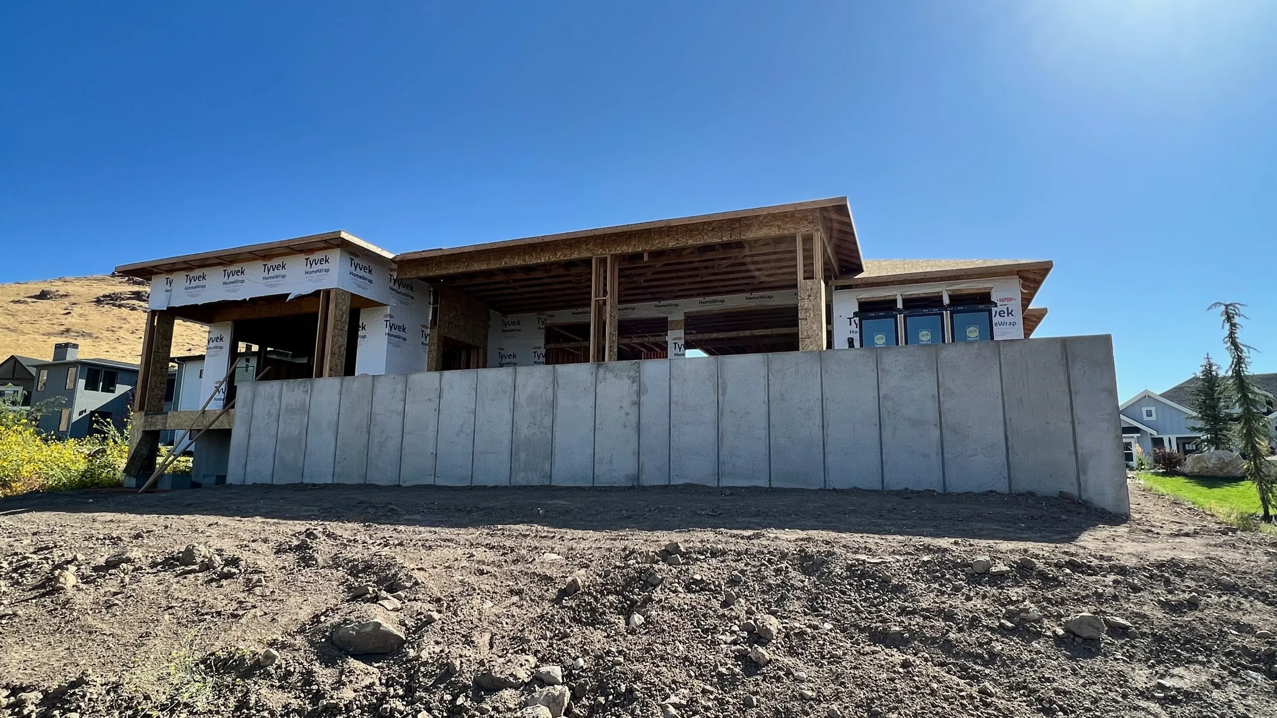 House under construction with a concrete foundation wall, wooden framing, and partially installed windows, set on a dirt lot with neighboring completed houses under a clear blue sky.