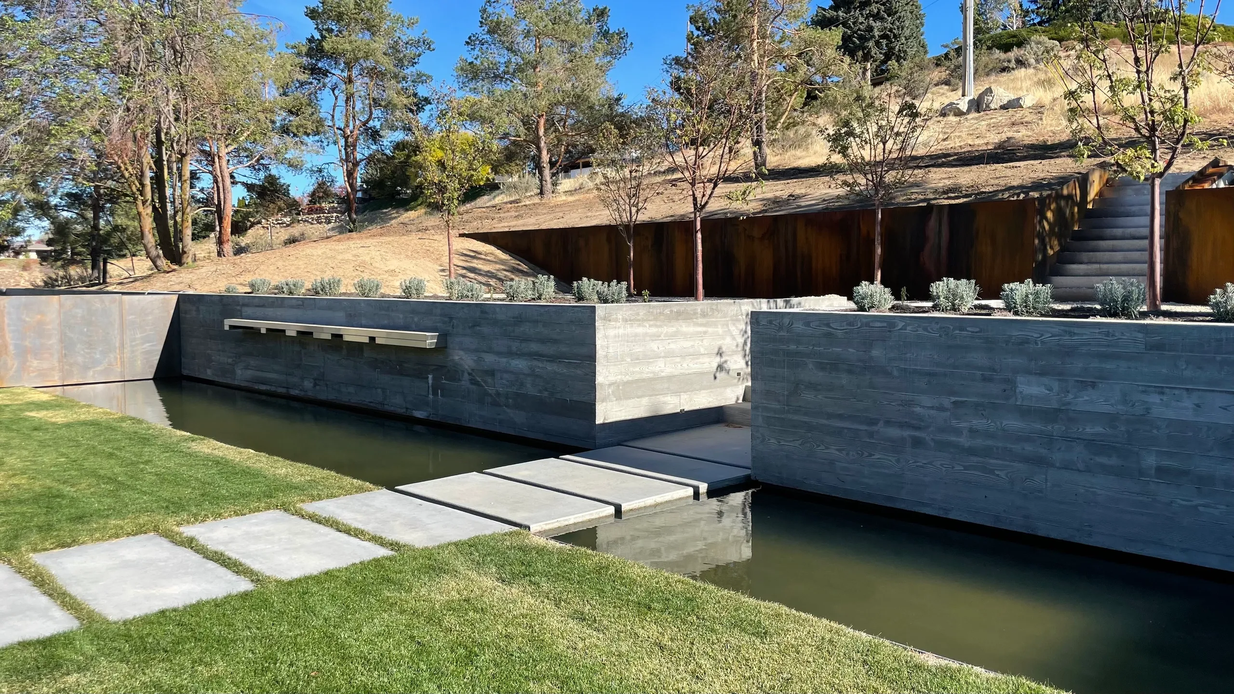 Modern outdoor water feature with concrete walls, a shallow canal, and stepping stones, surrounded by a grassy area and small trees on a sunny day.