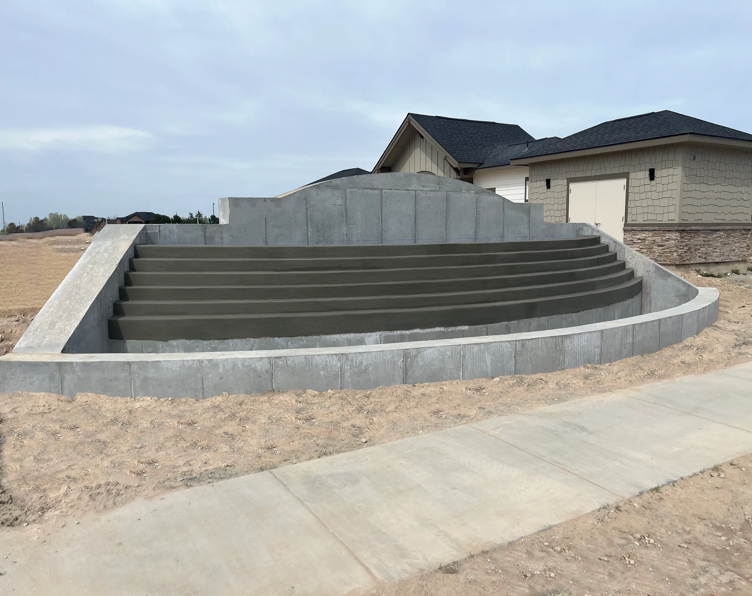 Concrete staircase with multiple steps, curved design, in front of a house with a gray and tan exterior, under an overcast sky, on a construction site or yard.