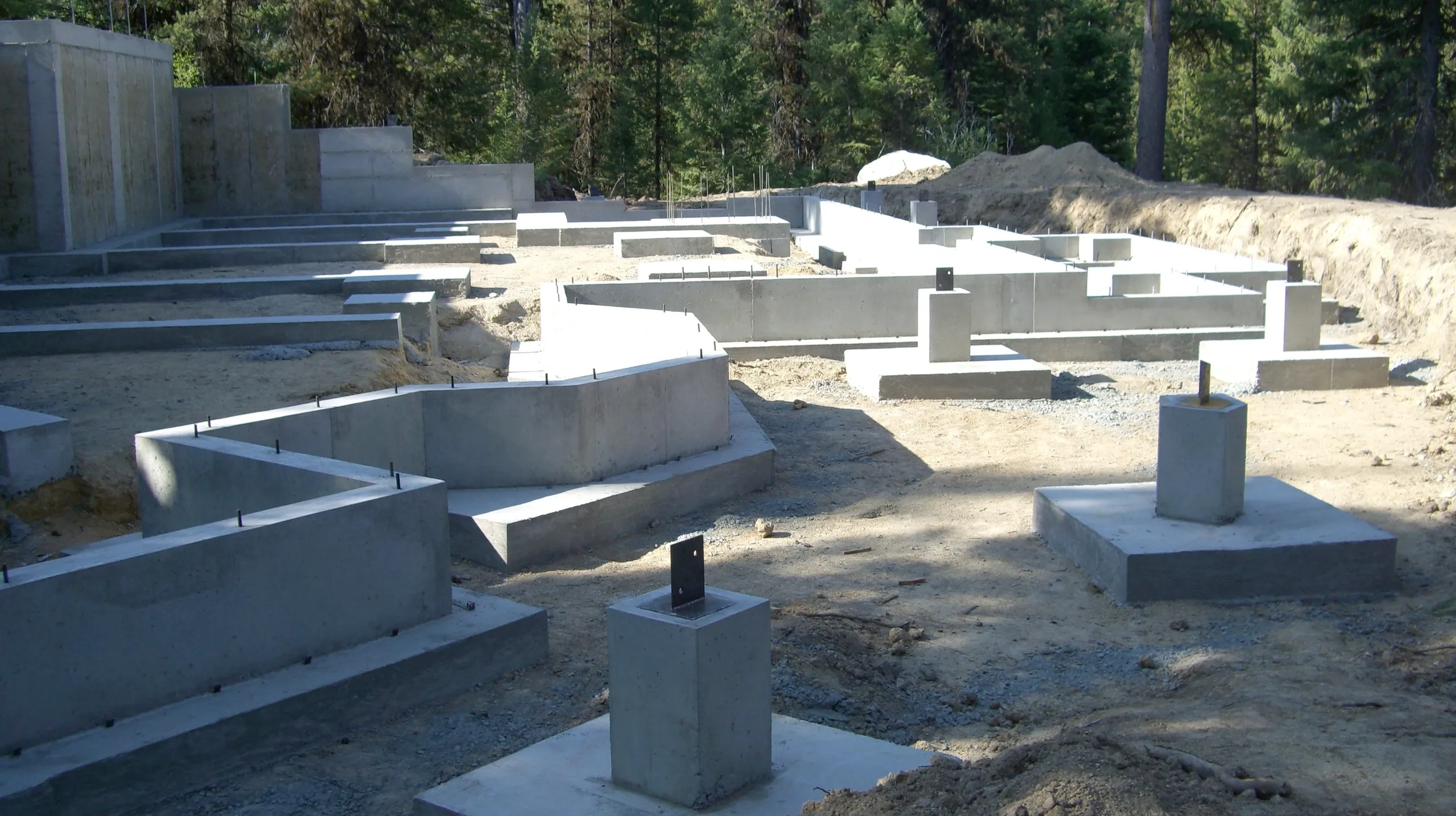 Foundation of a building under construction with concrete footings and rebar, surrounded by a forested area.
