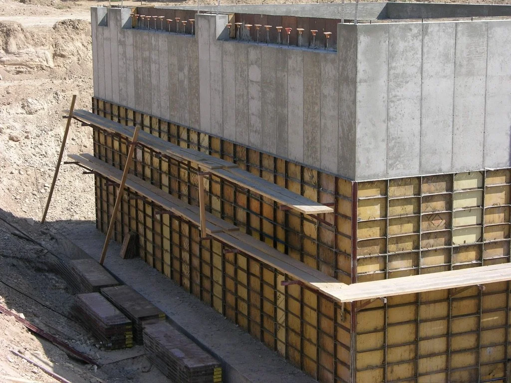 The image shows a construction site with a concrete building foundation with formwork in place for pouring the next concrete wall, and some wooden planks and construction tools nearby.