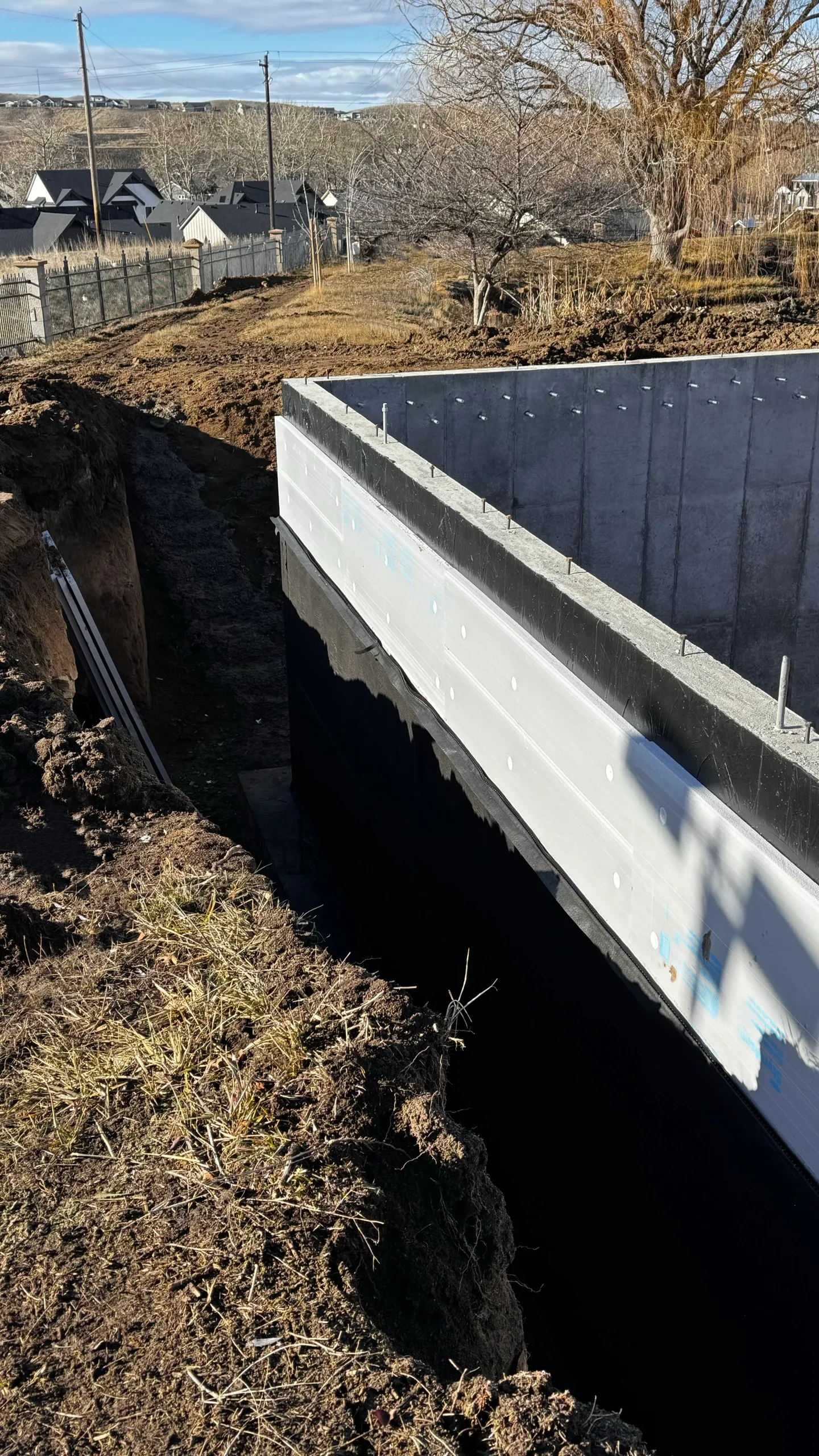 Construction site with an excavated trench and a newly installed concrete foundation for a building. Nearby trees and residential houses are visible in the background.