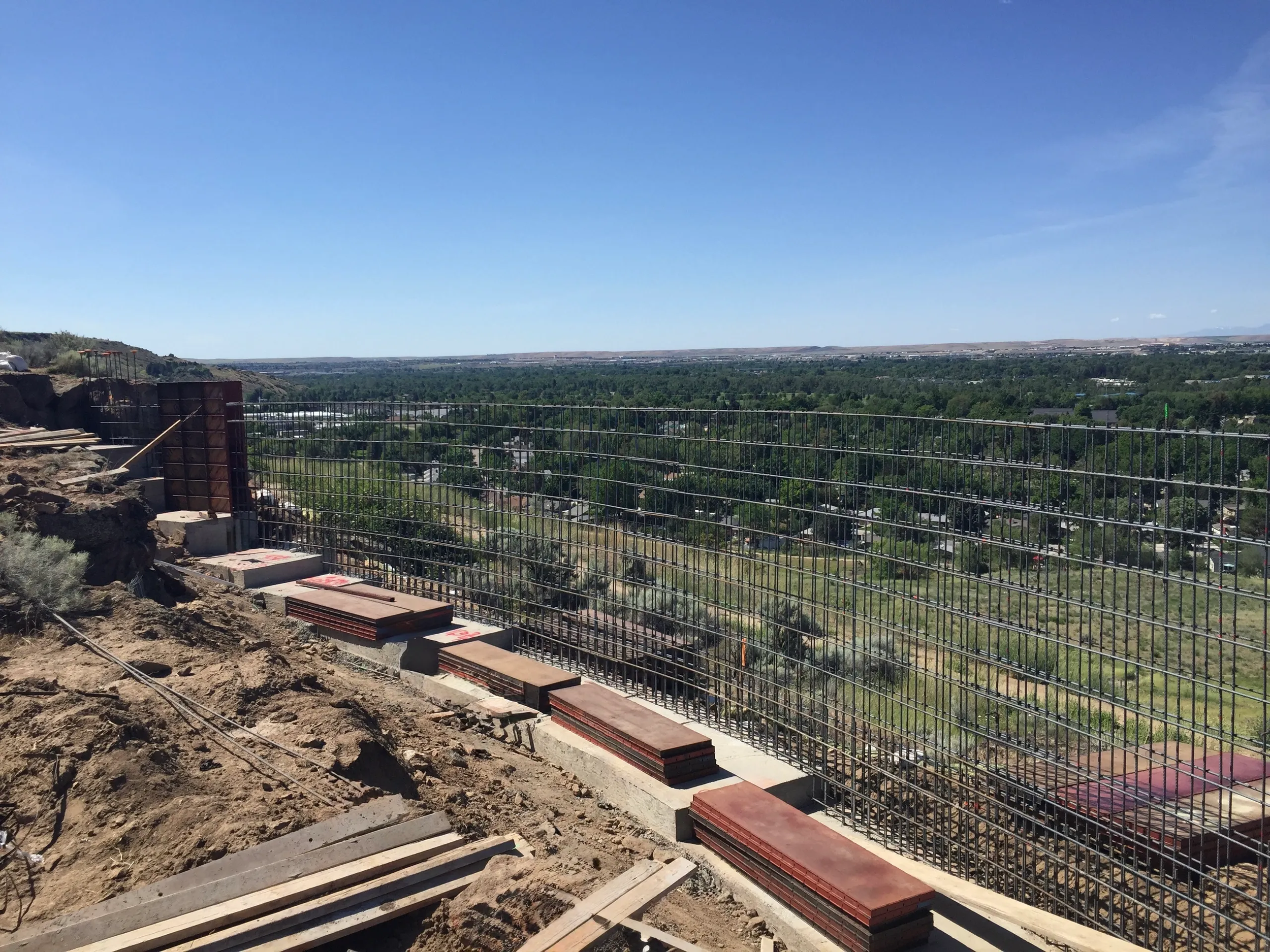 Construction site on a hill with a metal safety fence, view of a town with trees and buildings under a clear blue sky.