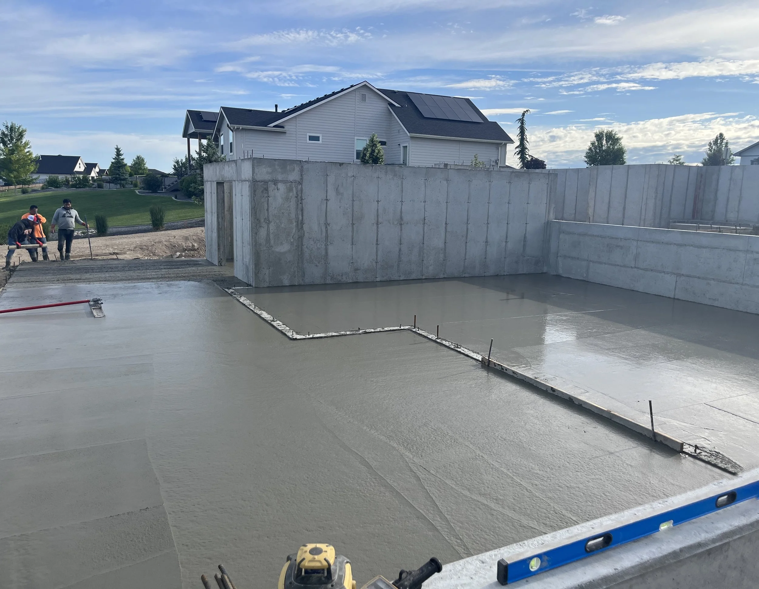 Construction workers pouring and smoothing concrete for a building foundation, with a retaining wall and residential houses in the background.