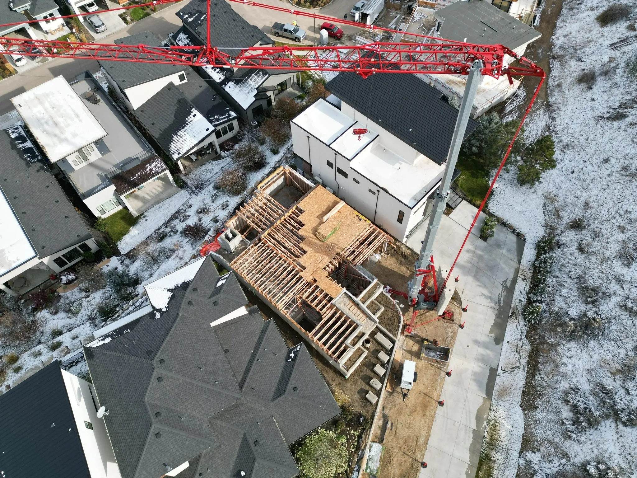 An aerial view of a house under construction, with a red crane lifting materials. Surrounding houses are visible along with snow on the roofs and ground.