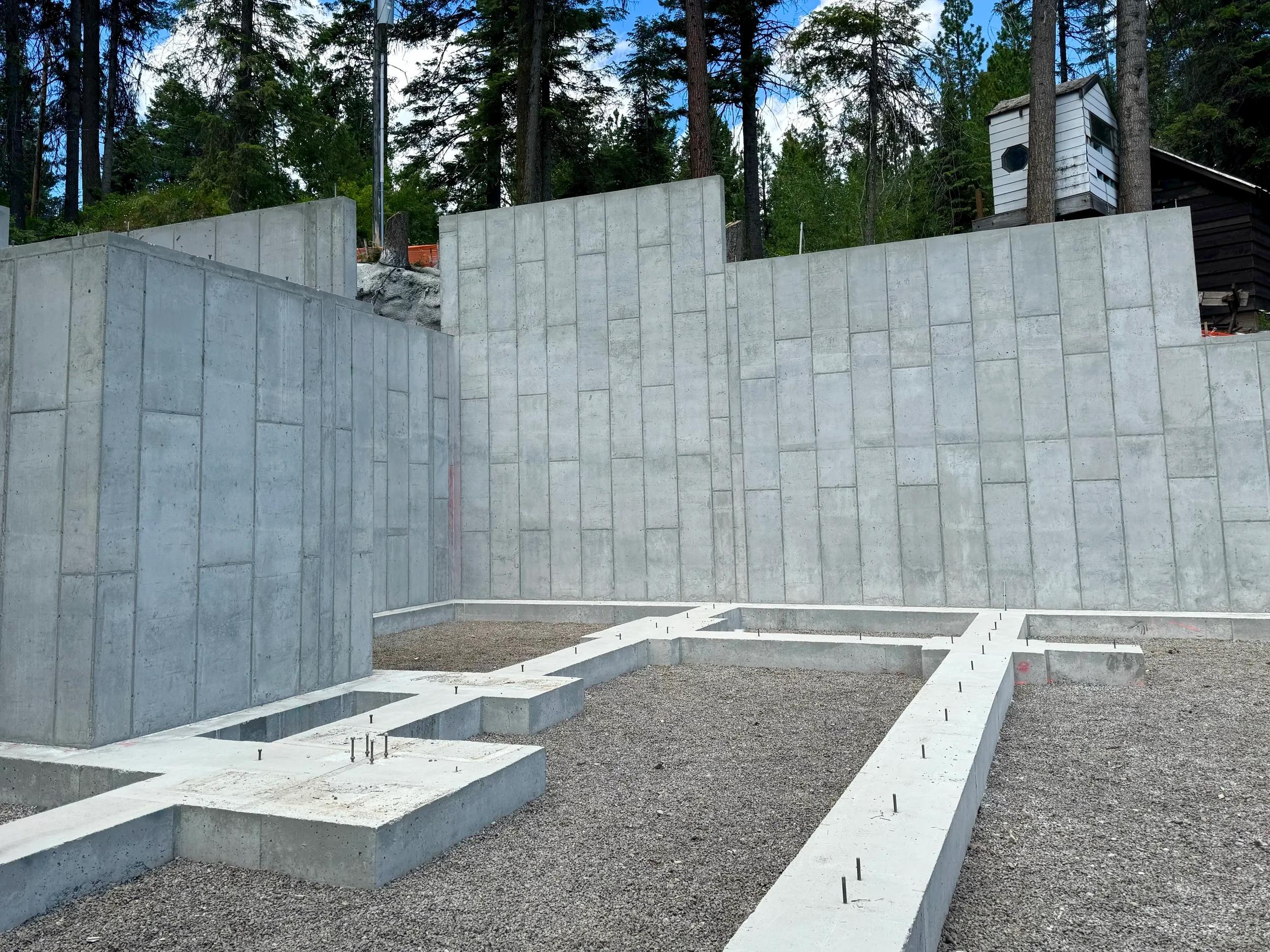 Construction site with concrete foundation and walls, surrounded by gravel, with trees and small structures in the background.