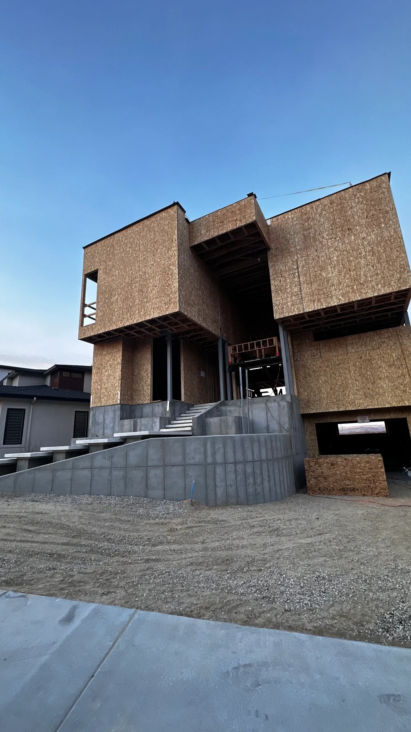 A modern, multi-story house under construction with unfinished wooden exterior walls and a concrete staircase leading to the entrance, set against a clear blue sky.