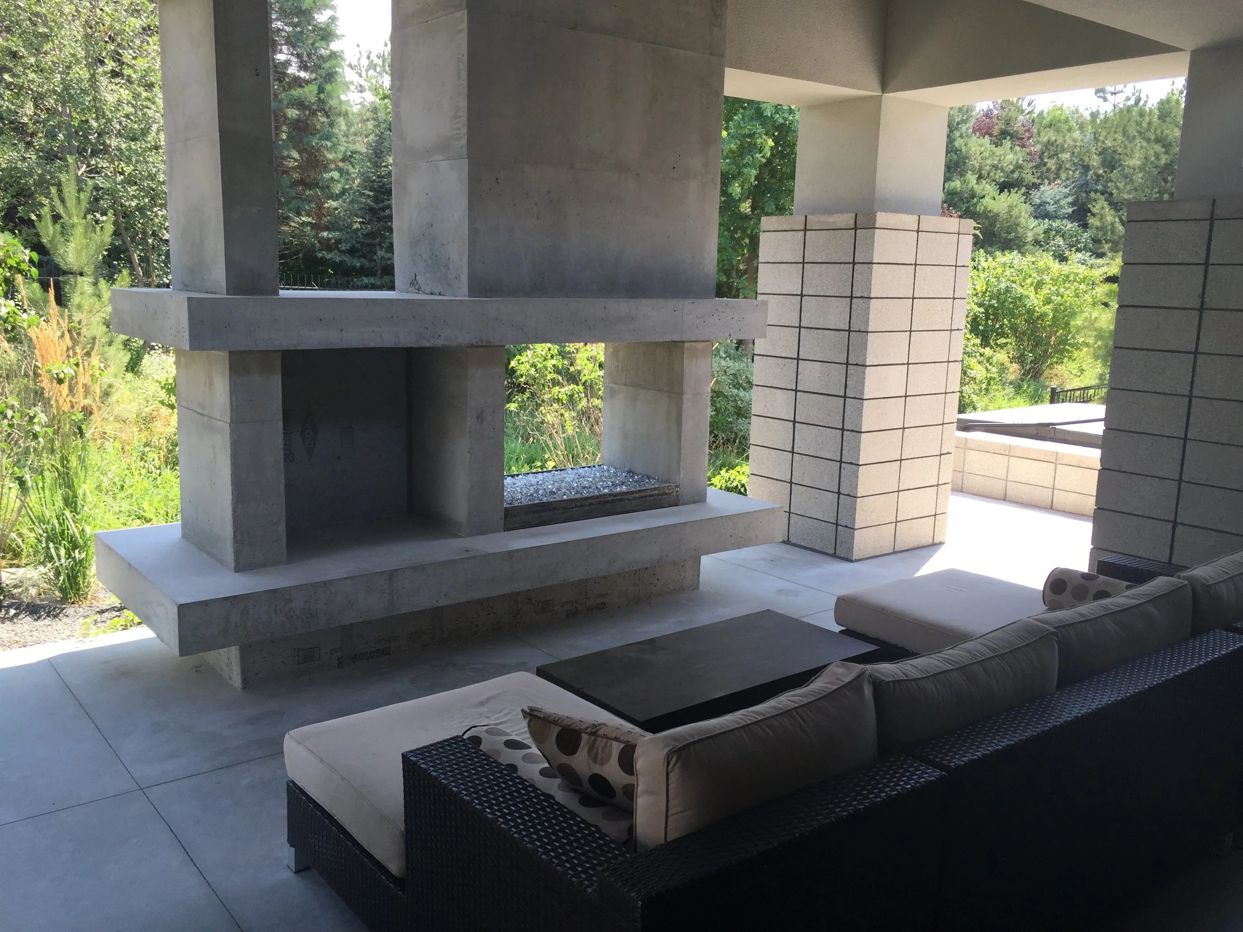 Modern outdoor living space with concrete fireplace, brick columns, and outdoor seating with cushions, overlooking lush greenery.