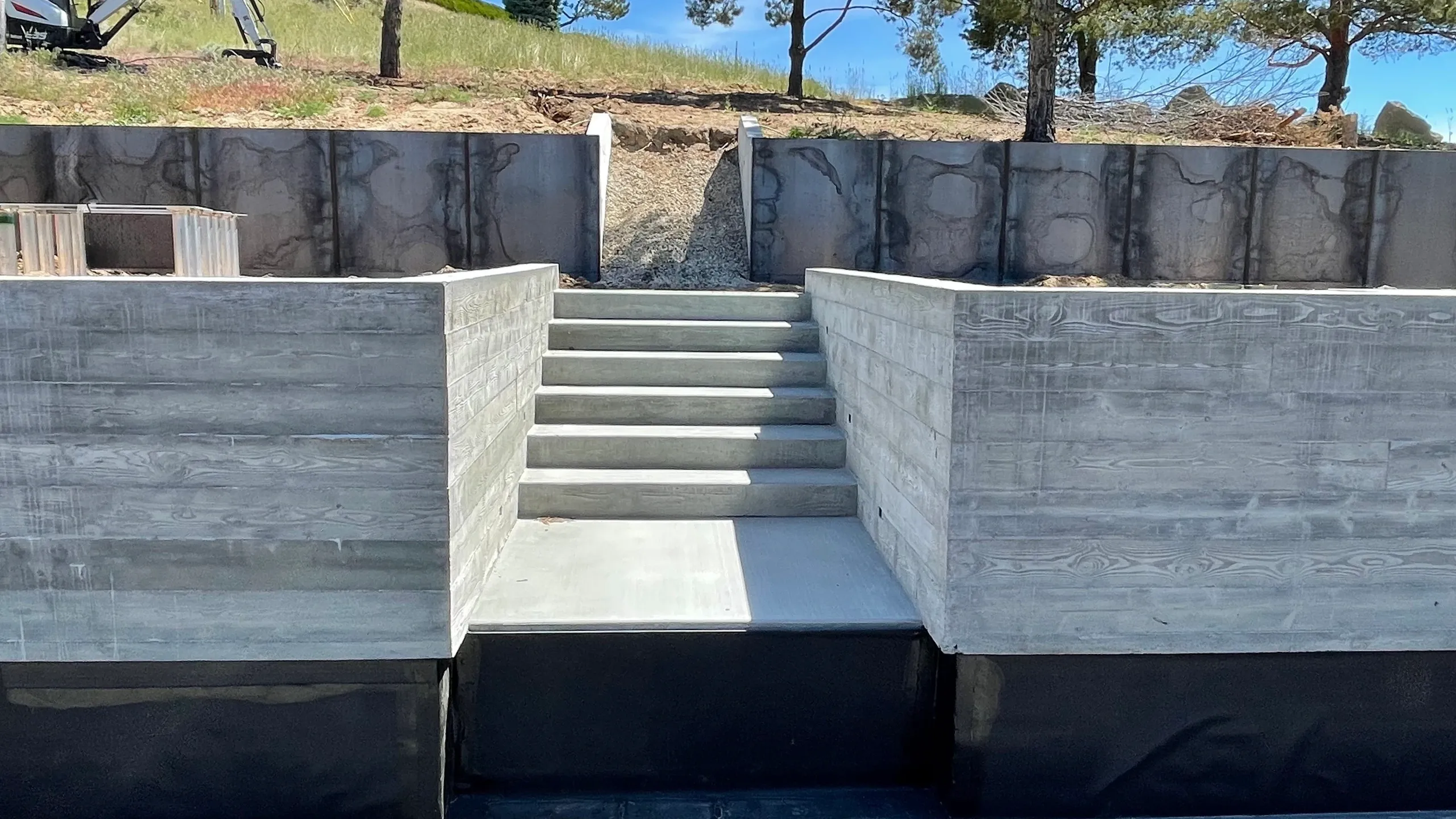 Concrete stairs leading up to a landscaped area with trees and rocks behind a concrete retaining wall.