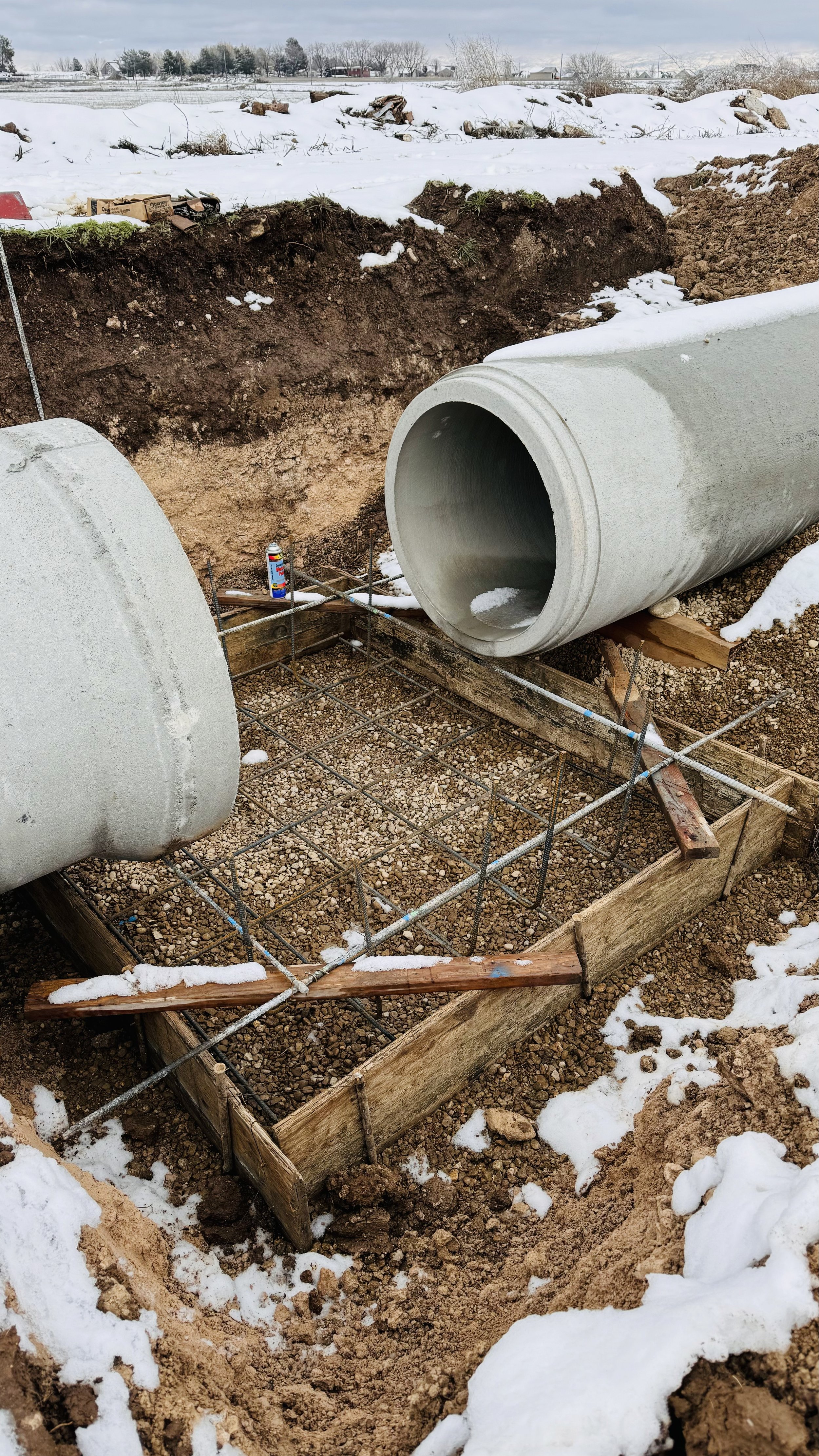 Construction site with large concrete pipes and rebar in a wooden formwork, snow-covered ground, and open field in the background.