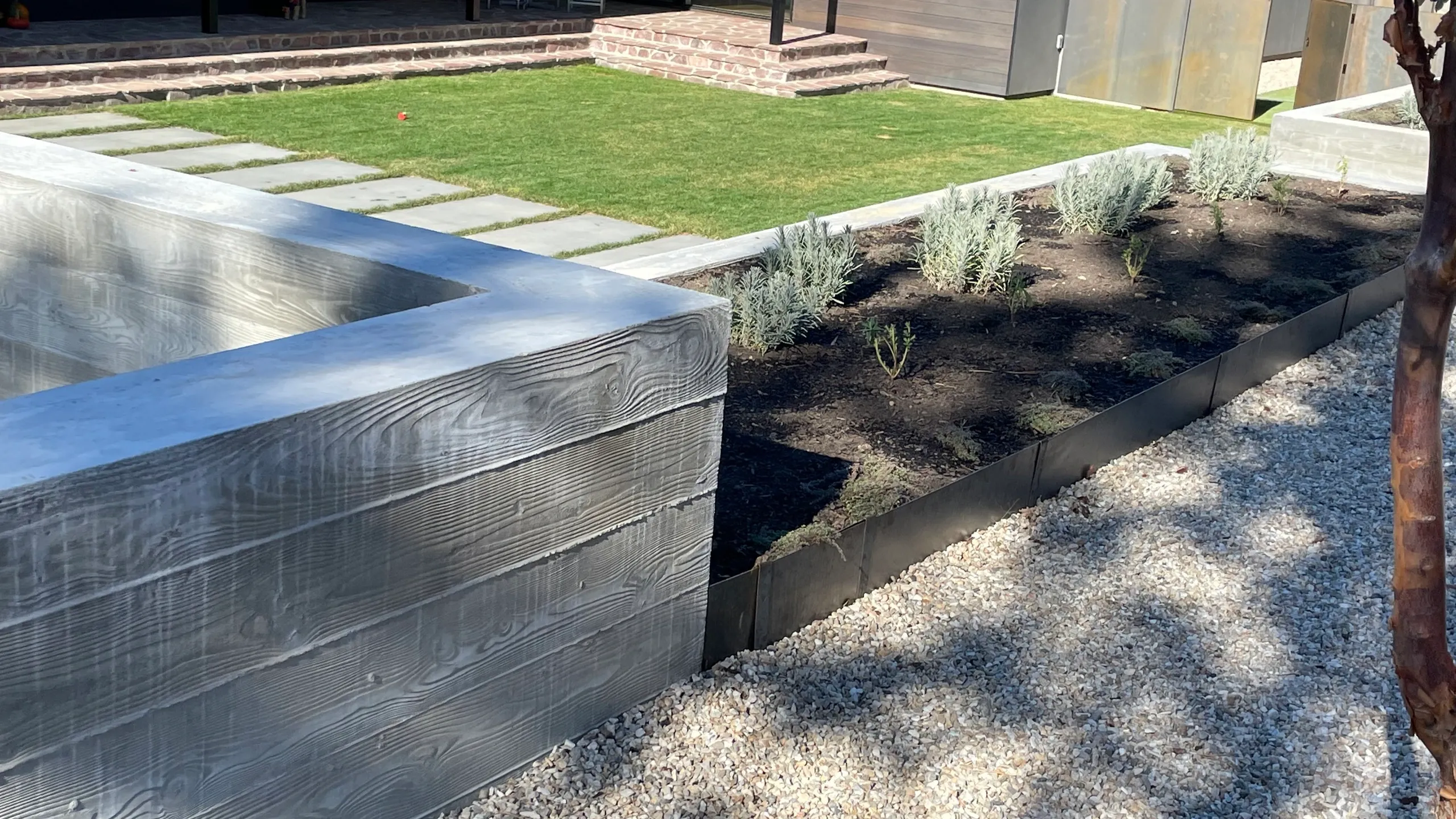 A landscaped yard with a walkway made of rectangular stepping stones, a patch of green grass, a flower bed with small plants, and a tree casting shadows on gravel.