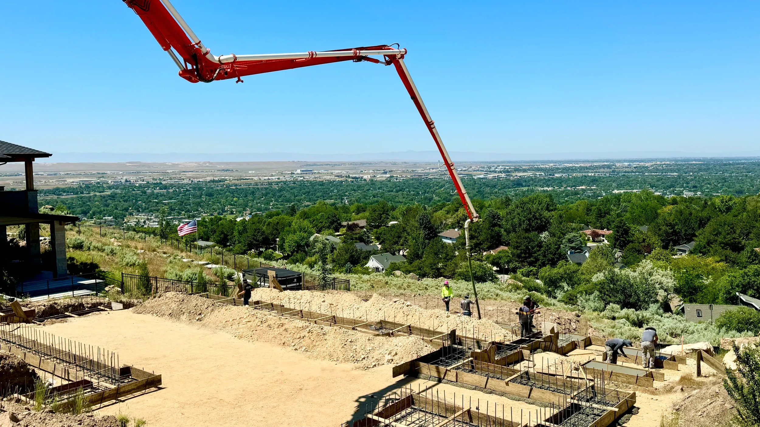Construction site on a hillside with workers, concrete forms, and a red concrete pump extending into the frame, overlooking a green landscape and distant cityscape under a clear blue sky.