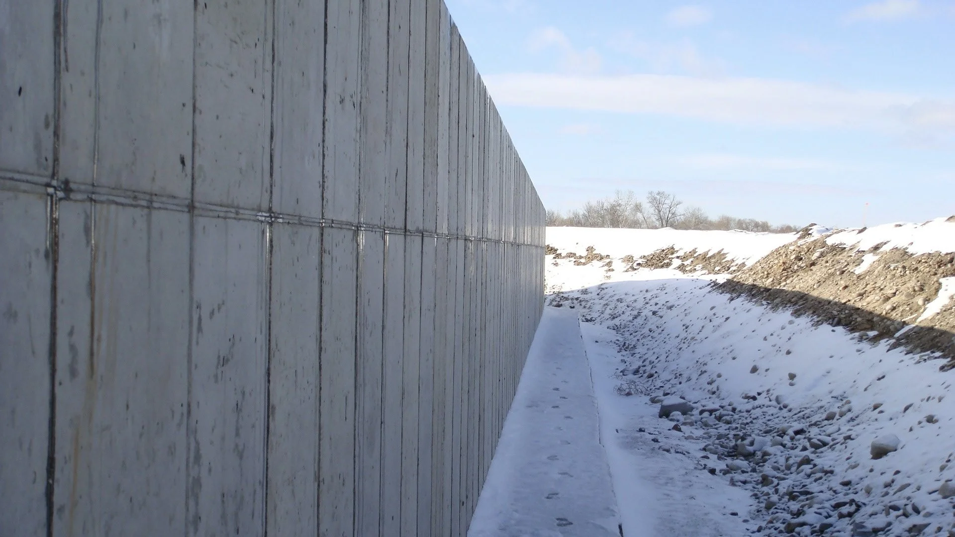A concrete wall on the left side of the image with snow and ice on the ground, and a clear blue sky in the background.